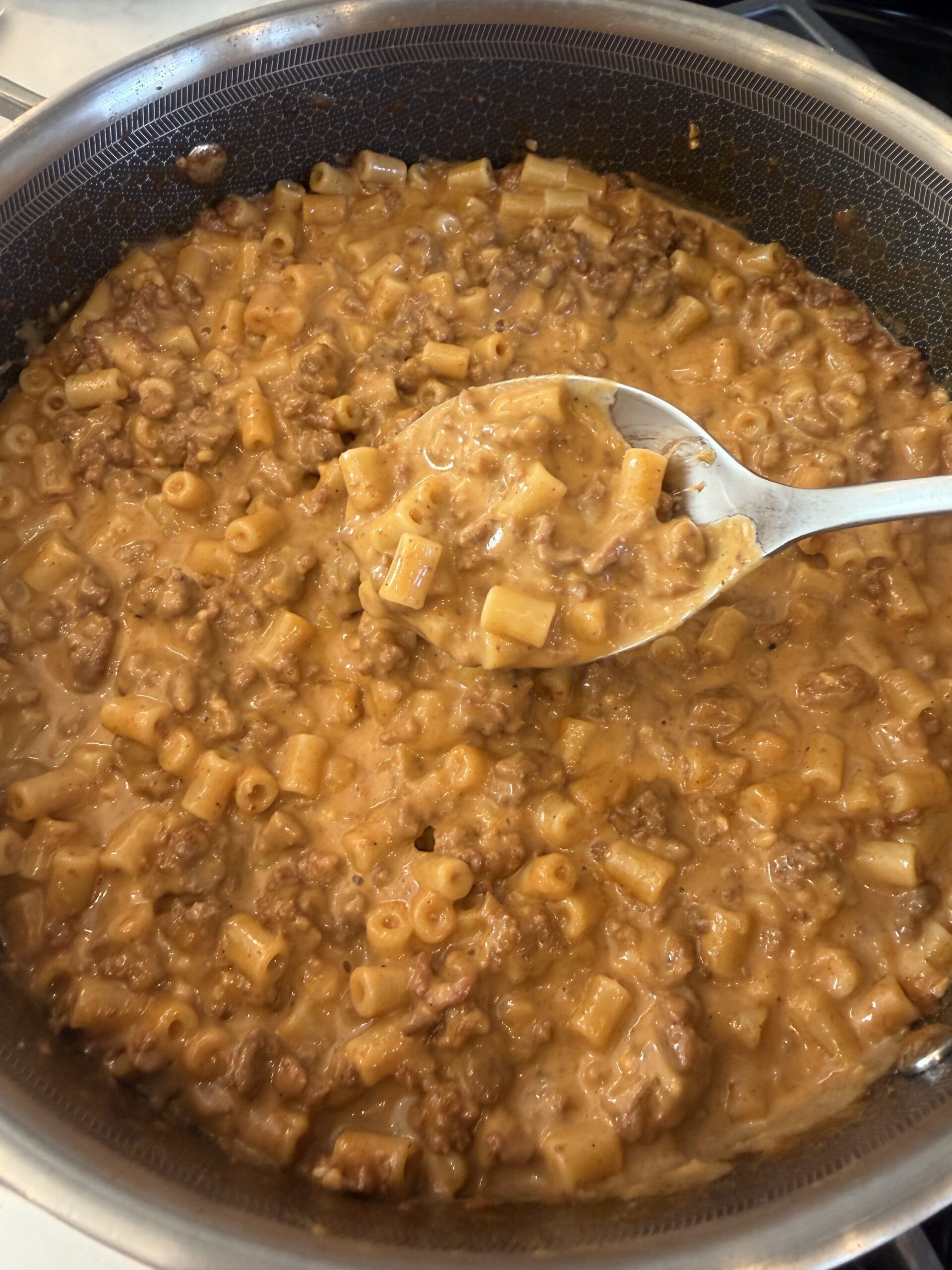 A close-up of a pot filled with creamy Homemade macaroni and ground beef, reminiscent of classic Hamburger Helper. A spoon is scooping up a portion, showcasing the cheesy, saucy texture of the dish.