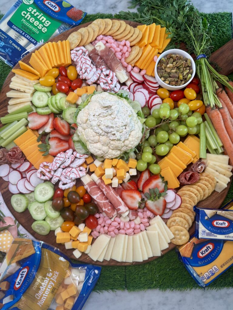 A charcuterie board with cheese, meats, crackers, veggies, fruits, nuts, and a cauliflower centerpiece. Kraft cheese packs and string cheese are on the sides, with herbs and carrots on a grass-like mat—a perfect display for any One Pot Momma gathering.