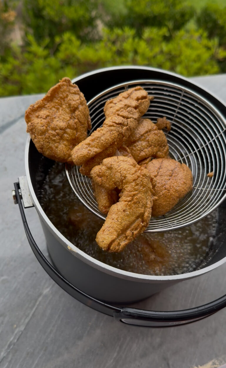 Four pieces of golden-brown fried chicken are lifted from hot oil in a black pot using a metal wire skimmer, reminiscent of preparing crispy fried fish. The background is slightly blurred with hints of greenery.