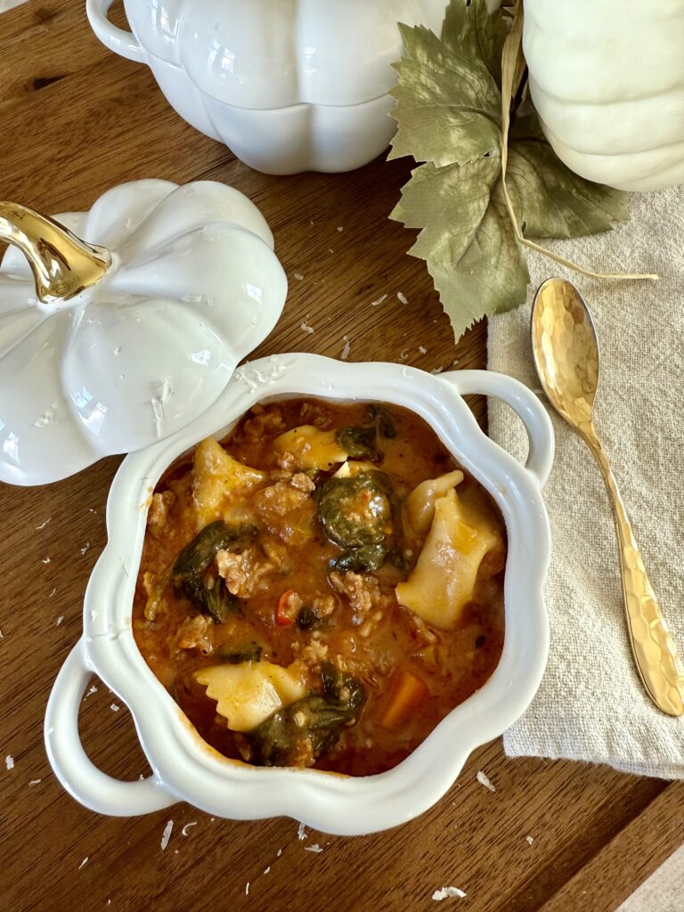 A white bowl of lasagna soup with spinach, ground sausage, and a hint of vodka sauce sits on a wooden table near a gold spoon, a white pumpkin-shaped dish, and decorative leaves.
