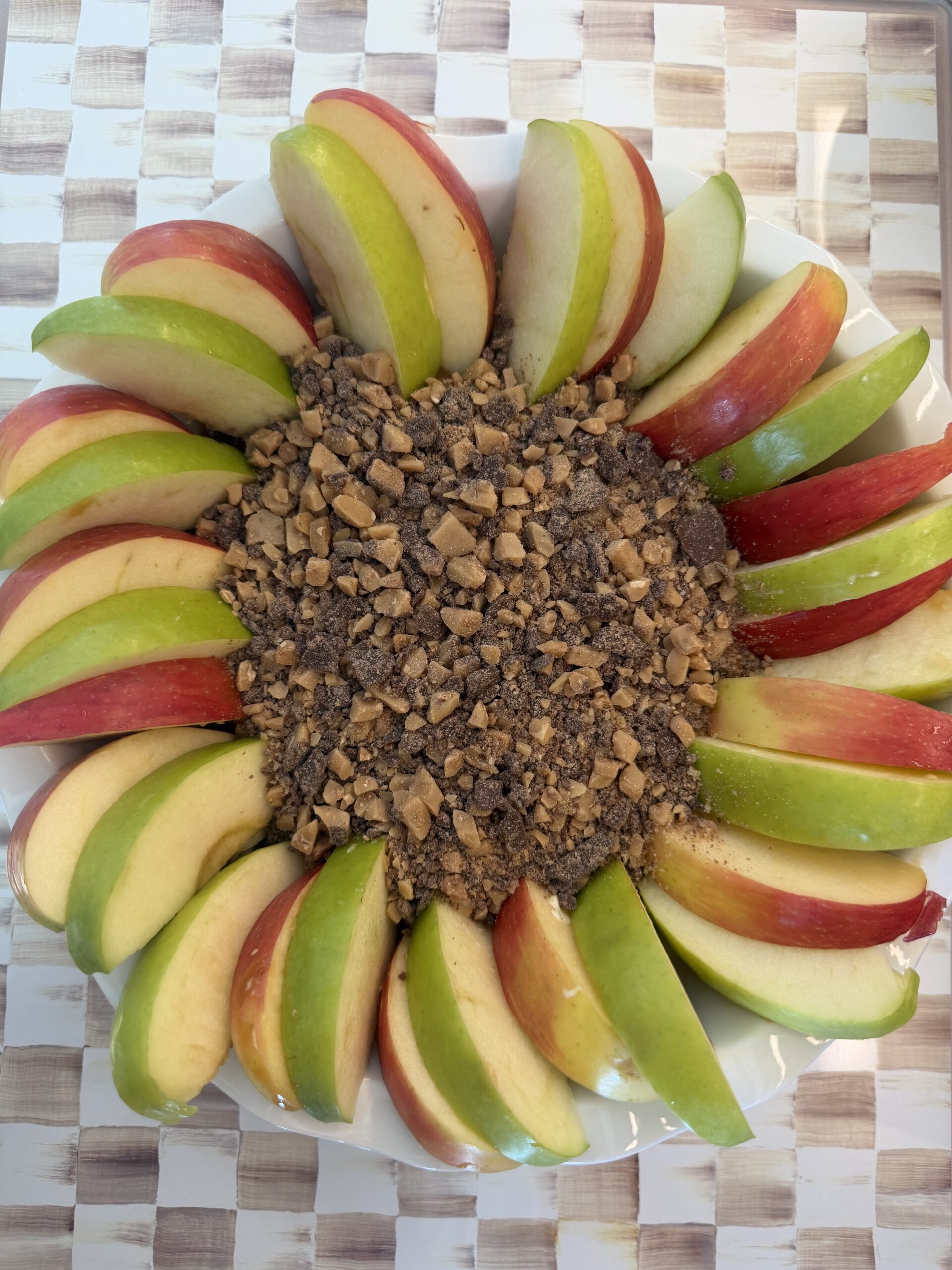 A plate of red and green apple slices arranged in a circle around a center pile of crushed toffee or chocolate candy pieces, perfect for dipping into your favorite Caramel Apple Dip, sits atop a checkered surface.