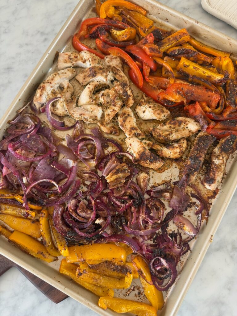Sheet pan with cooked strips of chicken, yellow and red bell peppers, and red onions, all seasoned for chicken fajitas and baked on a metal tray, resting on a marble countertop.