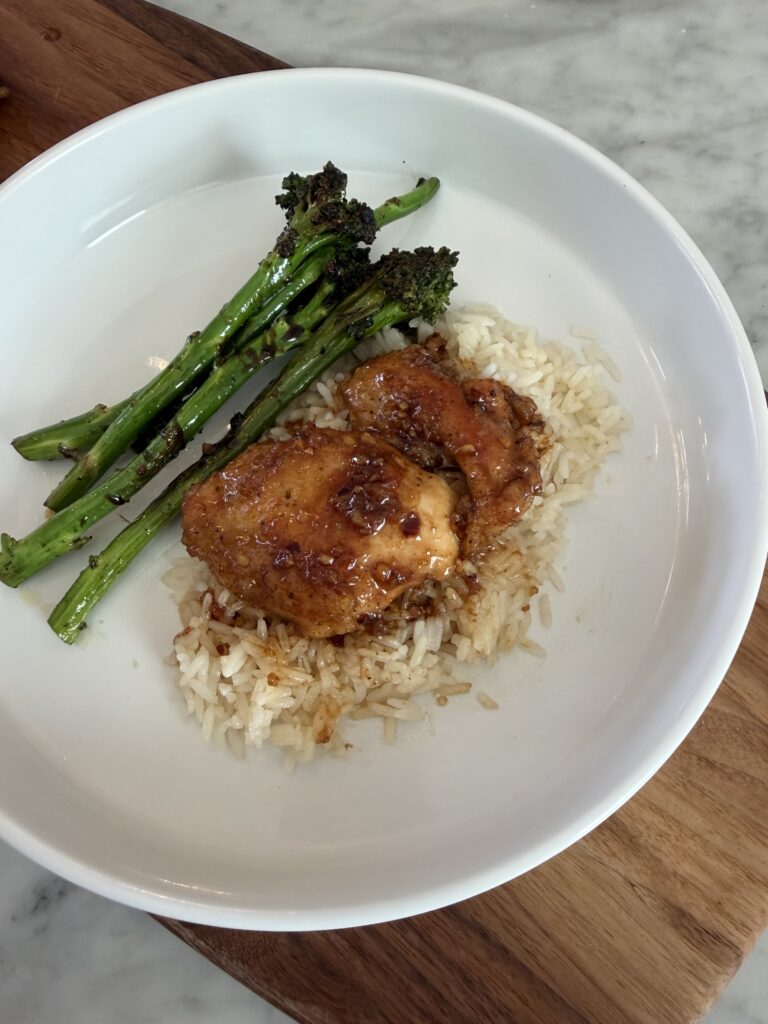 A white bowl with rice is topped with tender Honey Glazed Chicken Thighs, served alongside sautƩed broccolini, on a marble countertop.