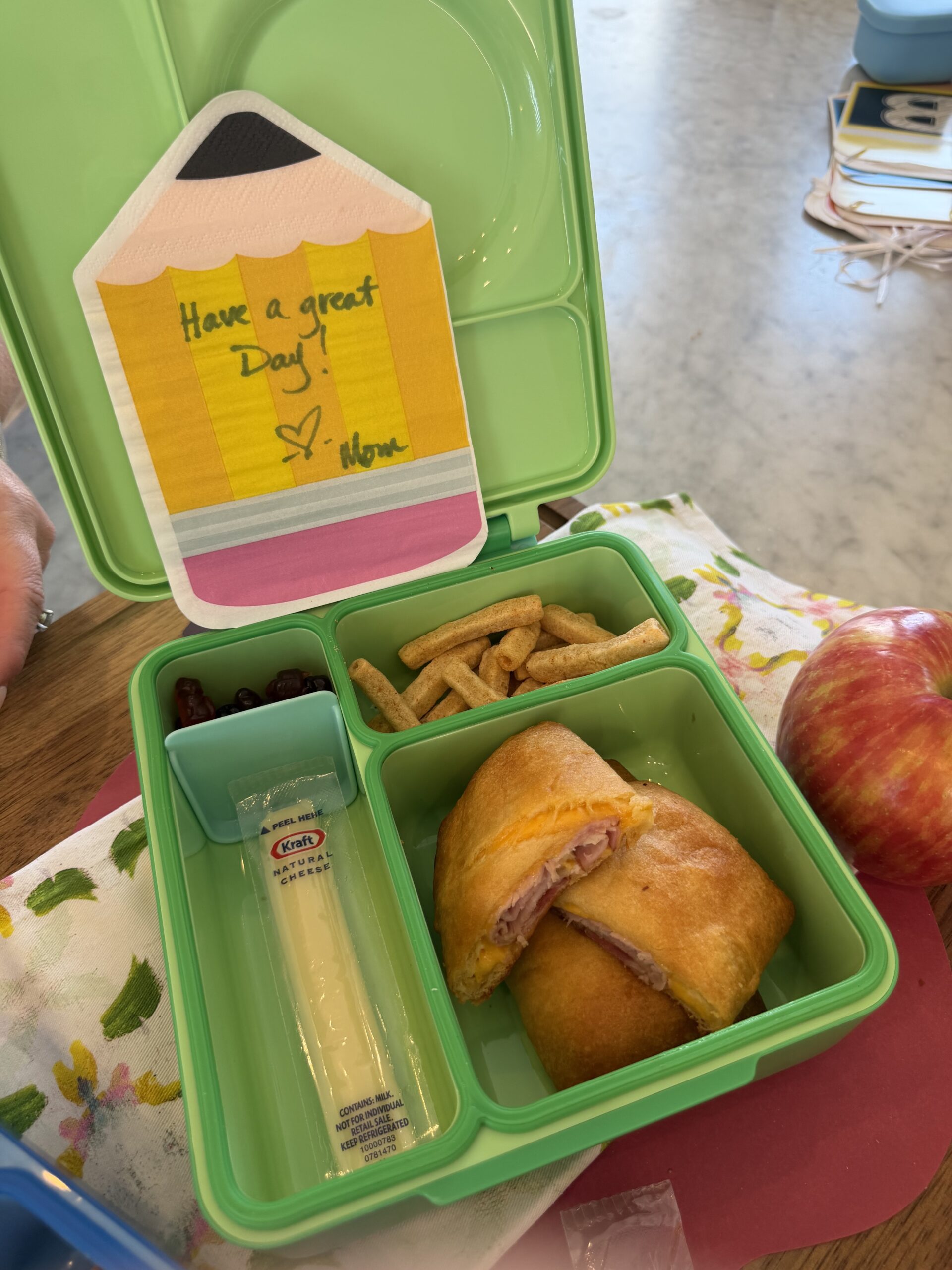 A green lunchbox with a ham and cheese croissant, string cheese, pretzel sticks, grapes, an apple, and a colorful note reading Have a great day! ♥ Mom. The lunch sits on a table with a floral napkin nearby.