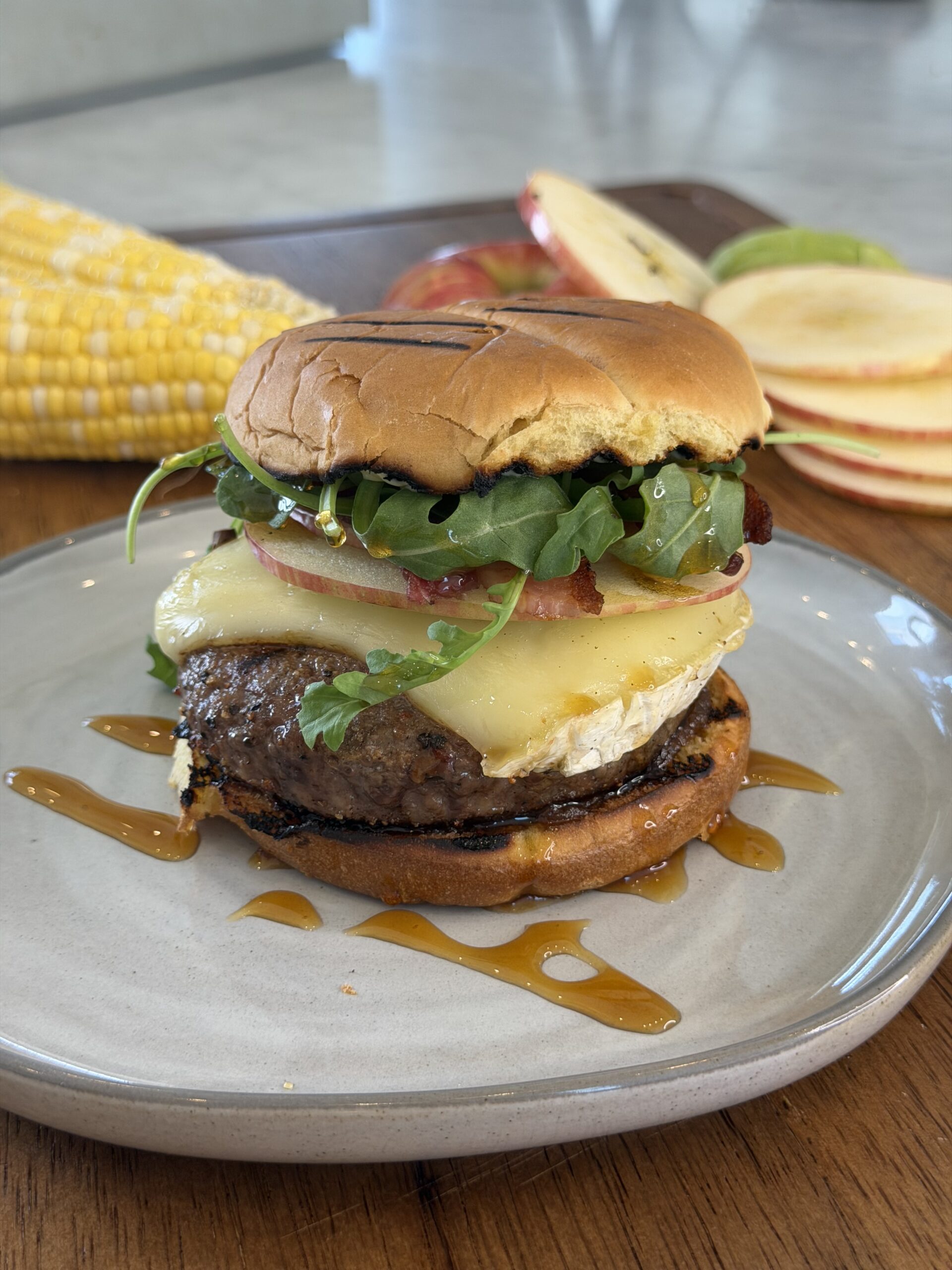 Experience our Apple Brie and Bacon Burger: a gourmet creation with crisp lettuce, creamy cheese, juicy tomato, and fresh arugula layered on a toasted bun and drizzled with savory sauce. Served on a plate with corn and apple slices in the background atop a wooden table.