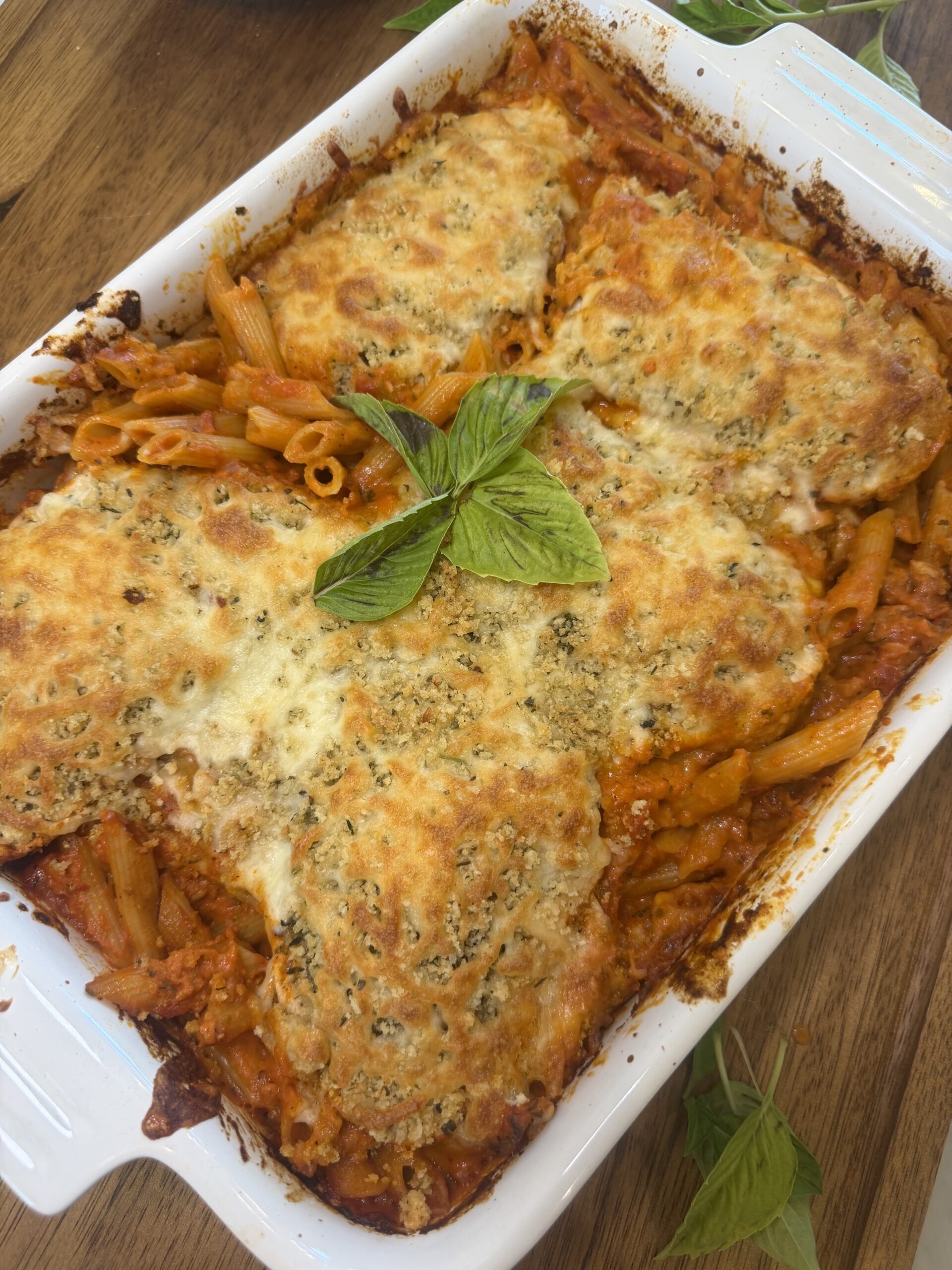 A white baking dish filled with penne pasta in tomato sauce, topped with melted cheese and herb-crusted chicken breasts, garnished with fresh basil leaves.