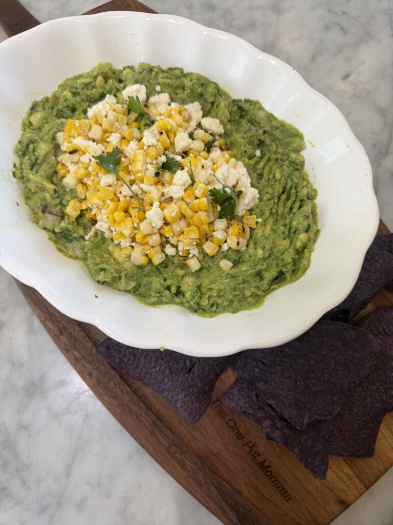 A white dish of guacamole, inspired by Mexican street corn, is topped with sweet corn, cheese, and cilantro. This flavorful Mexican appetizer is served on a wooden board with blue corn tortilla chips on the side.