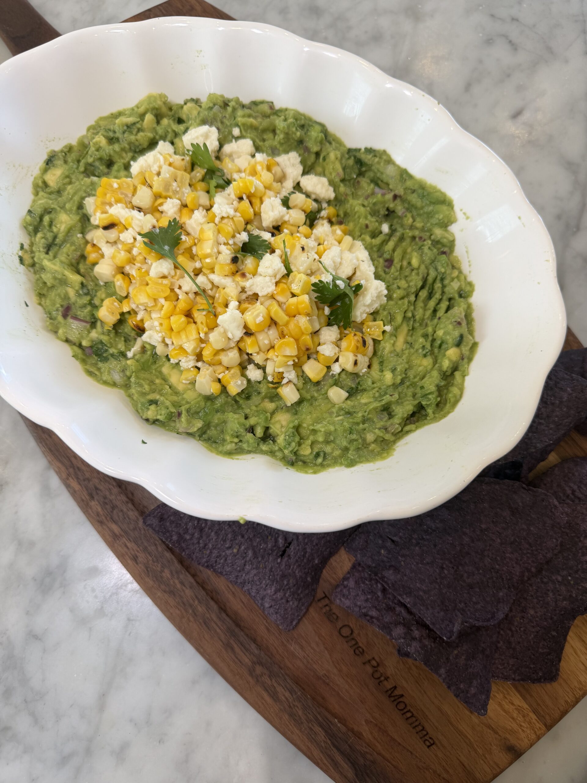 A white dish of guacamole, inspired by Mexican street corn, is topped with sweet corn, cheese, and cilantro. This flavorful Mexican appetizer is served on a wooden board with blue corn tortilla chips on the side.