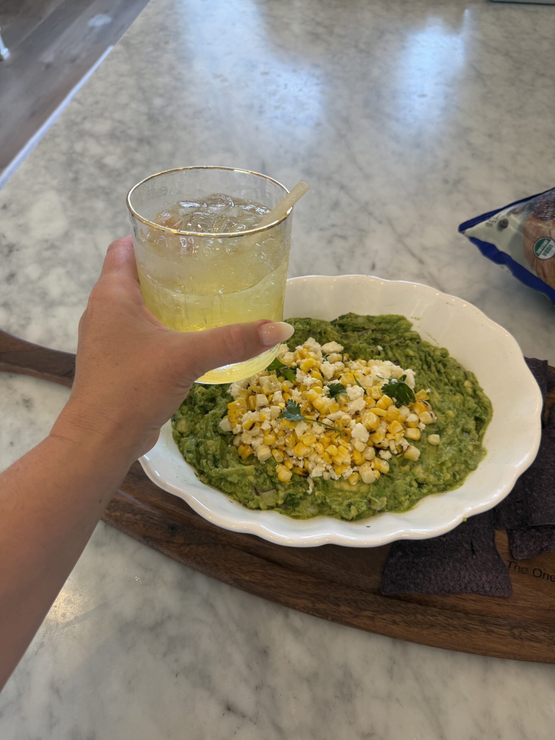 A hand holds a drink with a straw above a bowl of Mexican Guacamole topped with corn and herbs, set on a wooden tray with blue tortilla chips in the corner.
