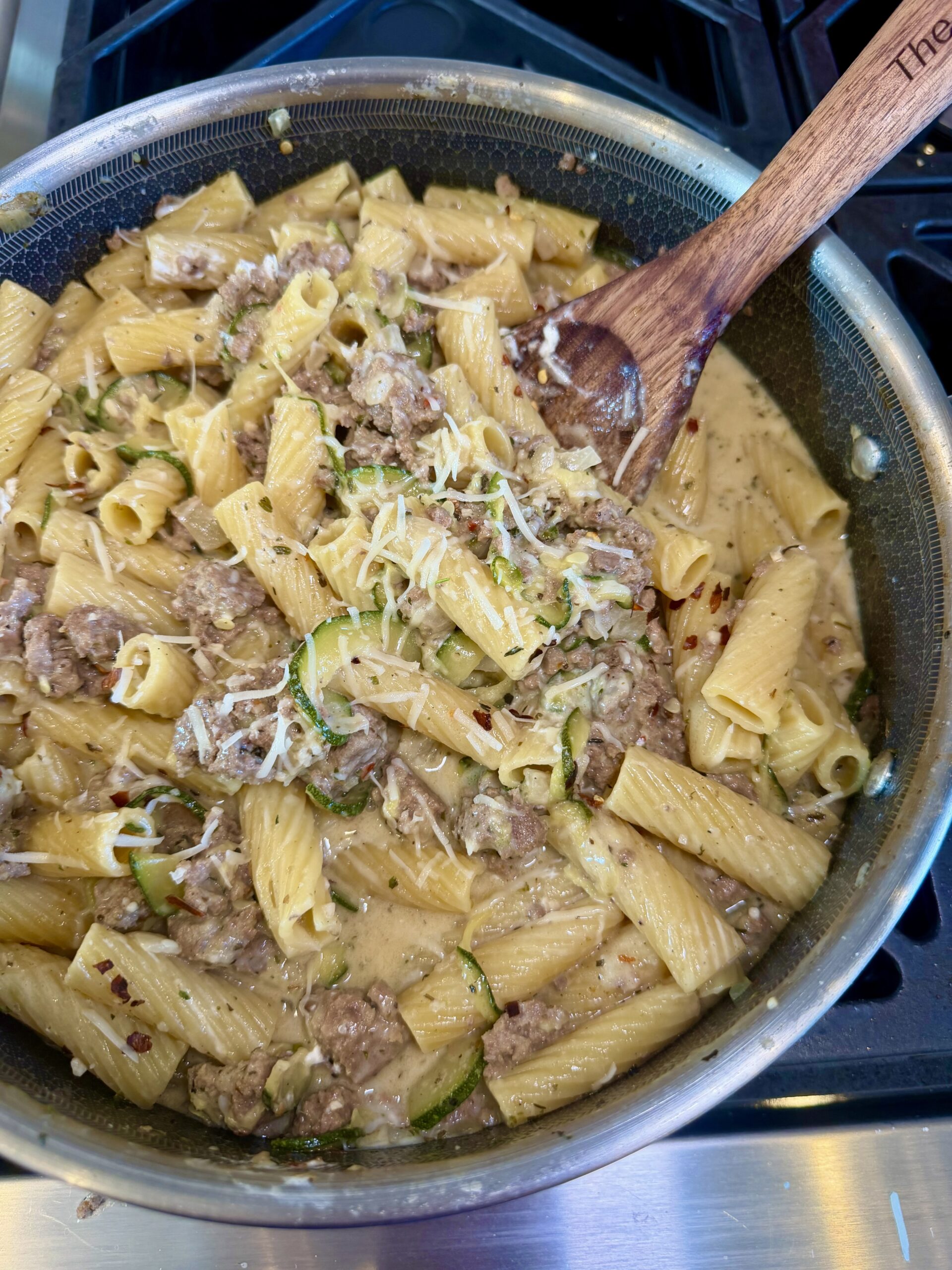 A skillet filled with creamy rigatoni pasta, ground meat, zucchini slices, and shredded cheese is being stirred with a wooden spoon on the stove—an easy and delicious One Pot Momma meal perfect for busy weeknights.