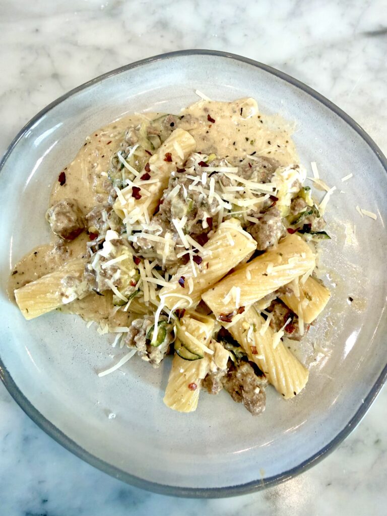 A plate of one-pot rigatoni pasta with a creamy garlic sauce, ground meat, zucchini, and shredded cheese, garnished with red pepper flakes on a marble surface.