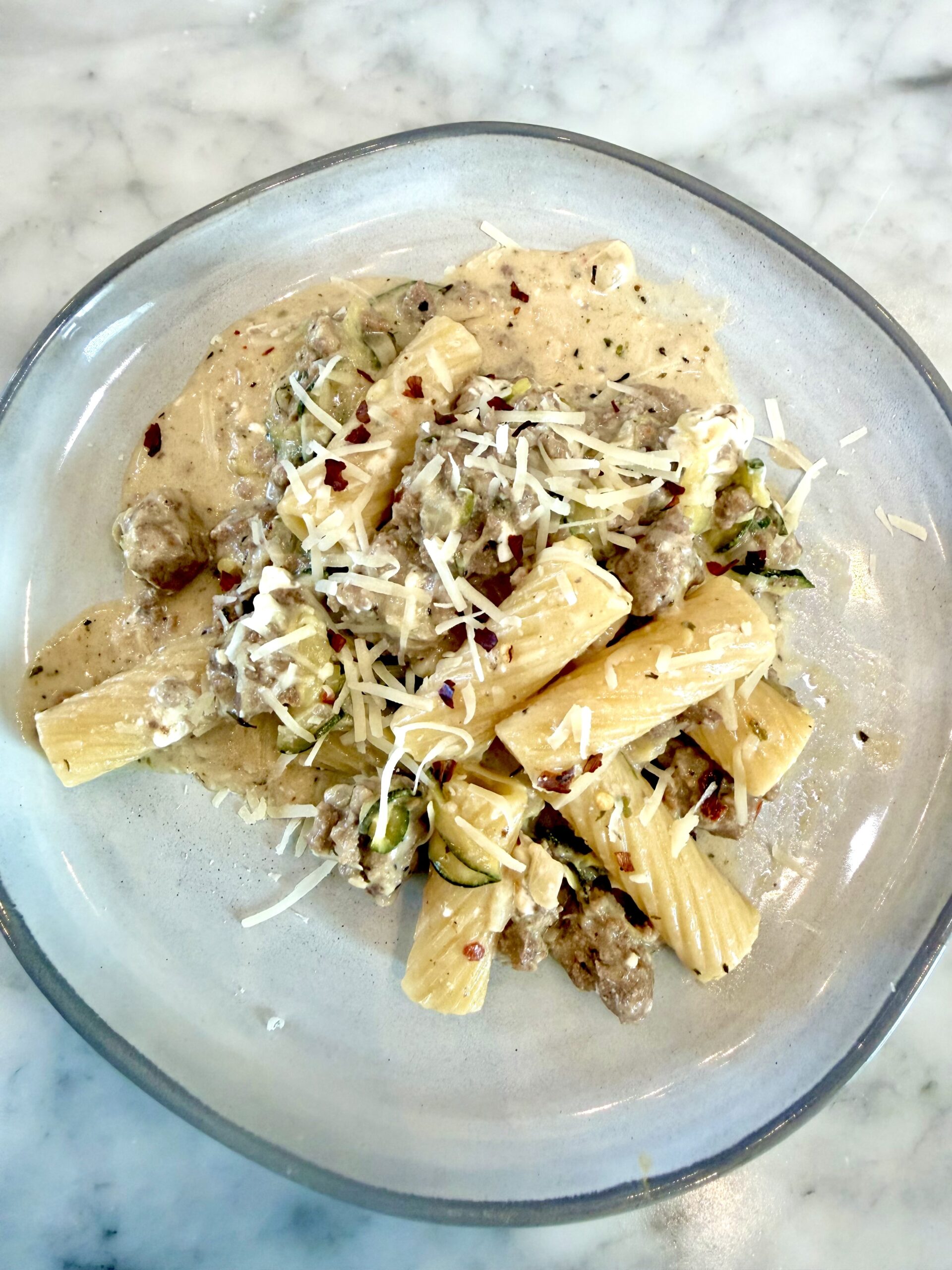 A plate of one-pot rigatoni pasta with a creamy garlic sauce, ground meat, zucchini, and shredded cheese, garnished with red pepper flakes on a marble surface.