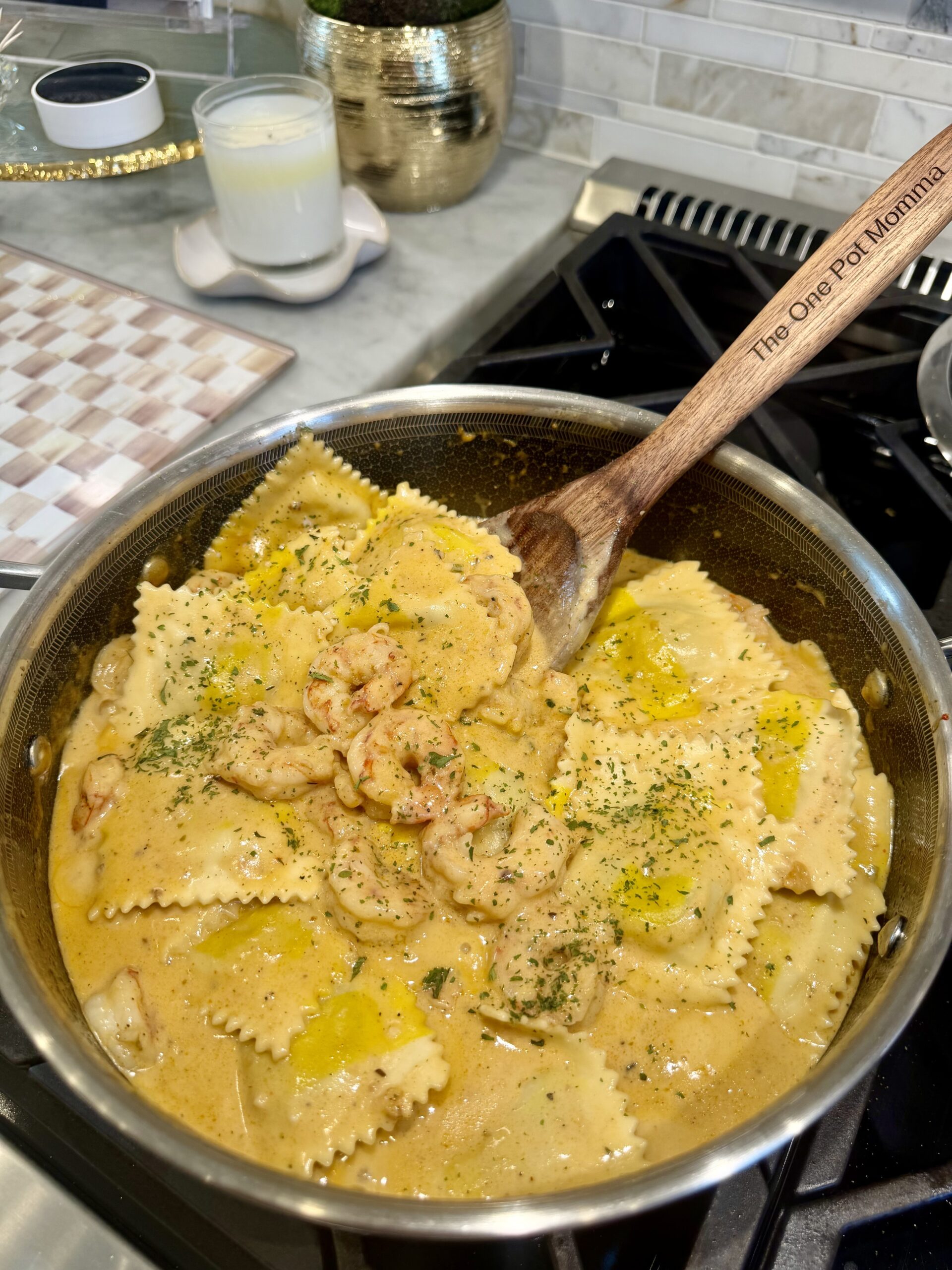 A skillet of creamy Lobster Bisque with Ravioli garnished with herbs sits on a stovetop, with a wooden spoon labeled The One Pot Momma stirring the dish. A candle and kitchen decor are visible in the background.