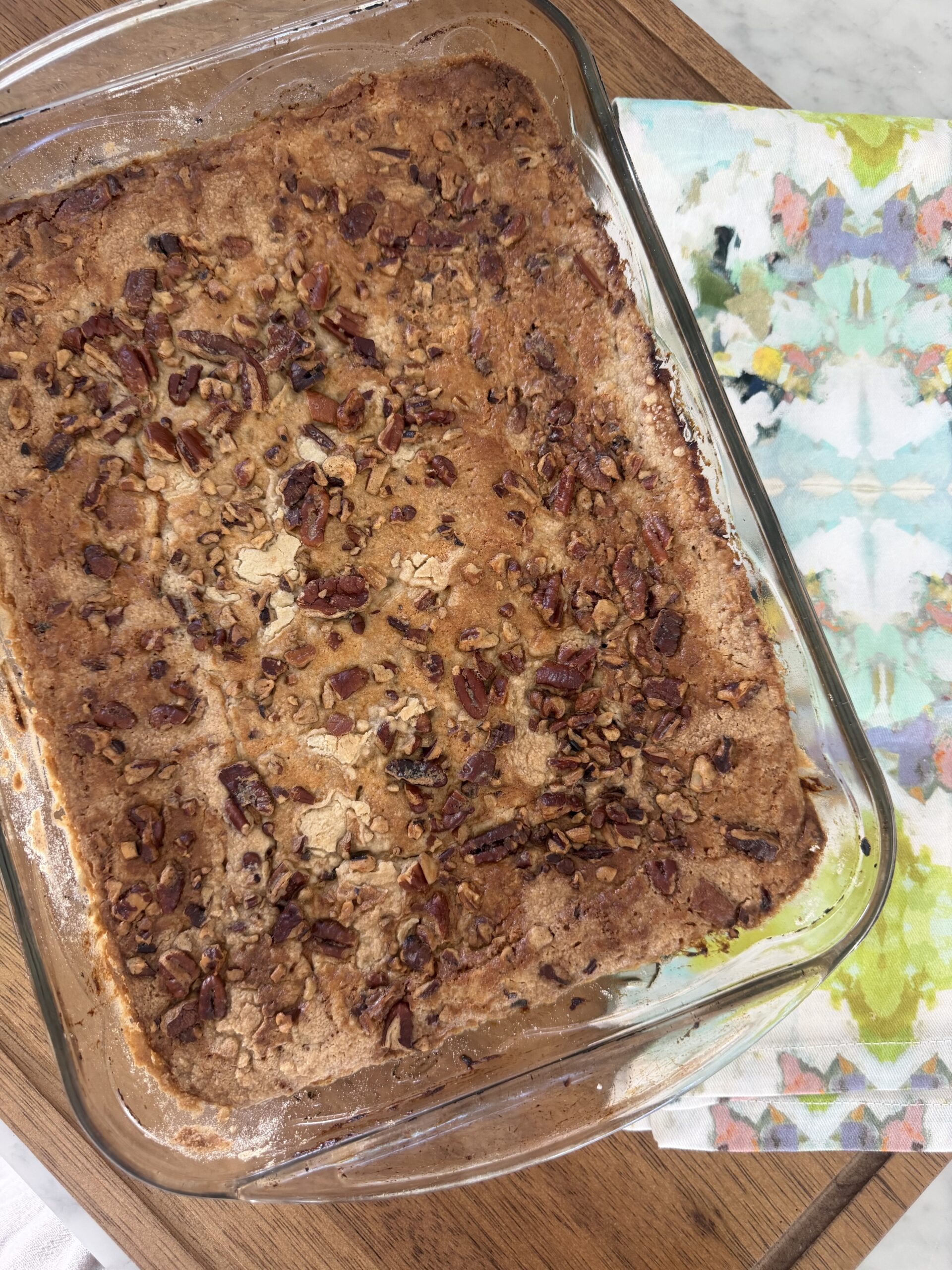 A glass baking dish with a golden-brown Maple Pecan dessert topped with chopped pecans sits on a wooden board next to a colorful floral napkin.