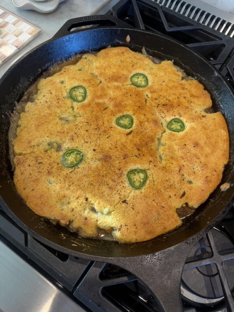 A golden brown cornbread topped with jalapeño slices is baked in a black cast iron skillet on a stove.