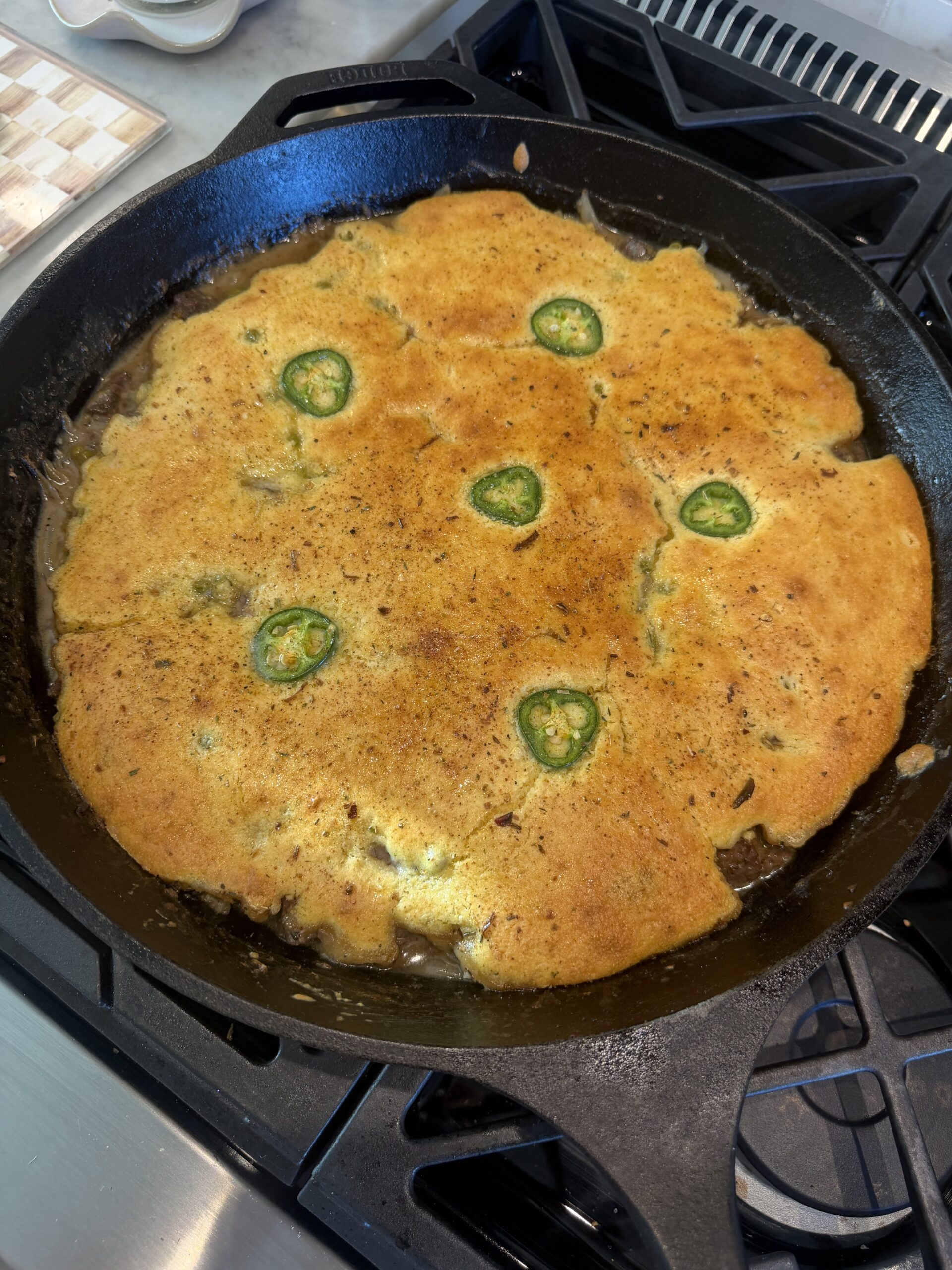A golden brown cornbread topped with jalapeño slices is baked in a black cast iron skillet on a stove.