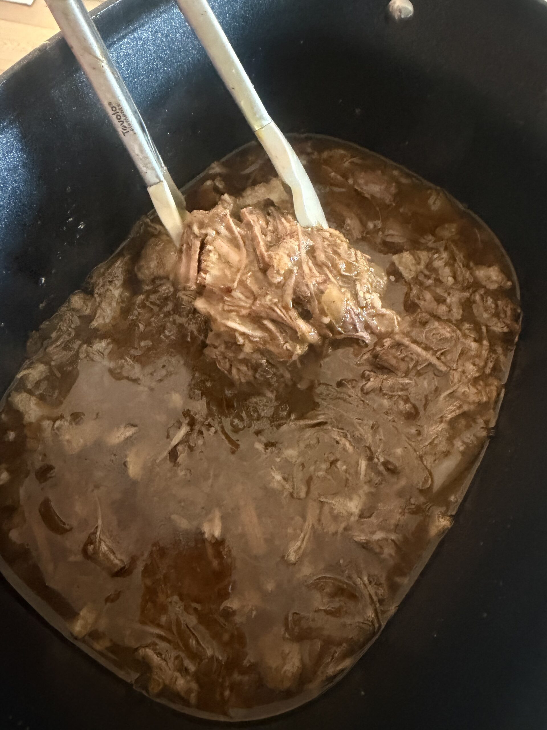 Shredded beef is lifted from a Crockpot of rich brown broth using metal tongs, with more tender beef and savory liquid visible in the pot.