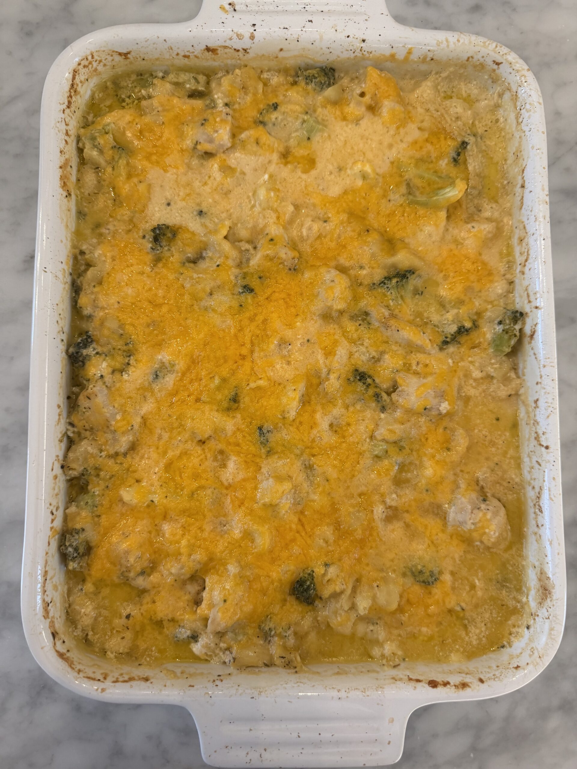 A baked casserole in a white dish, topped with melted cheese and containing pieces of broccoli and chicken in a creamy sauce, placed on a marble countertop.