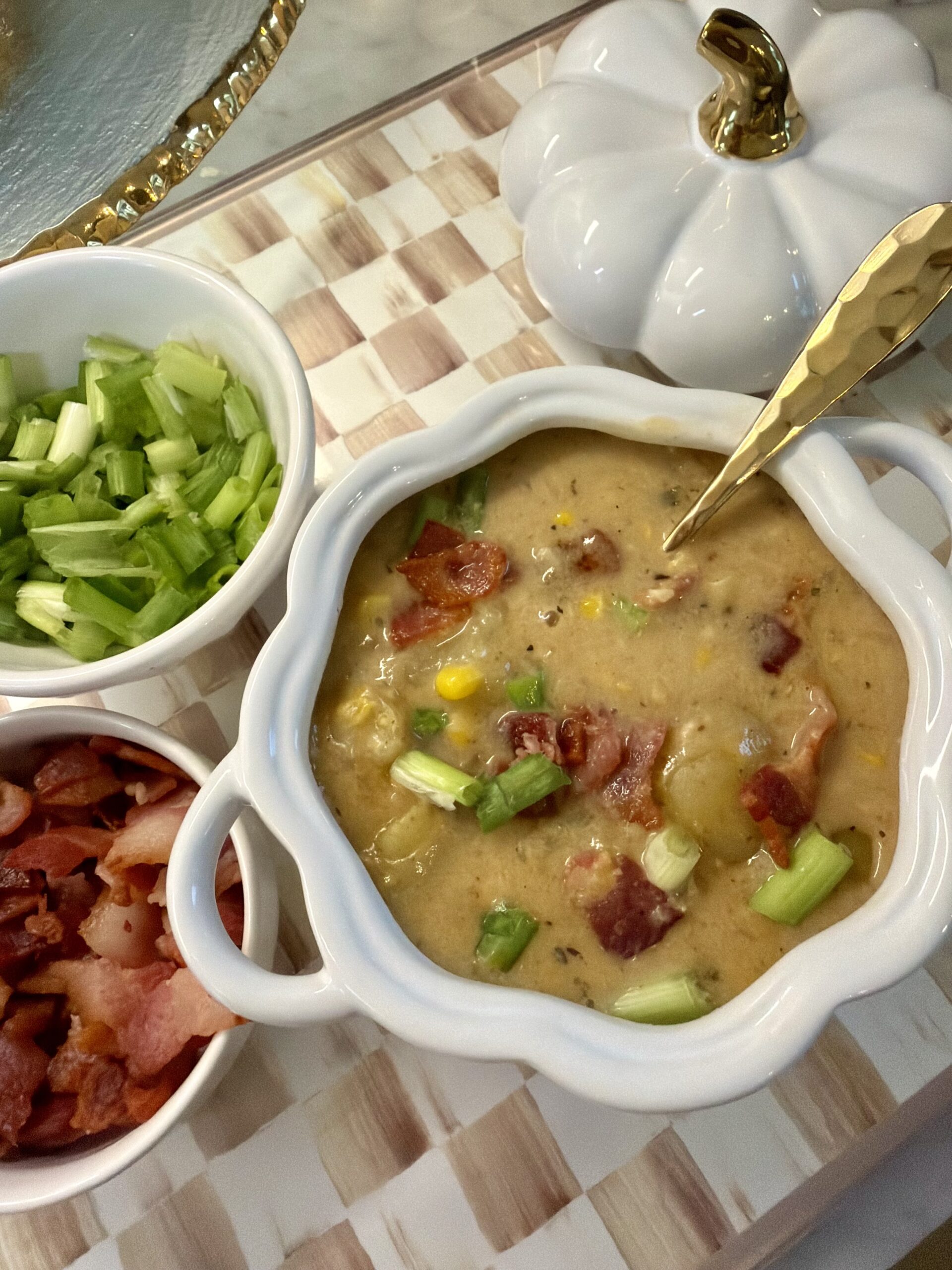 A bowl of creamy soup with bacon, corn, and green onions sits next to small bowls of chopped green onions and bacon on a tray, with a white pumpkin-shaped container nearby.
