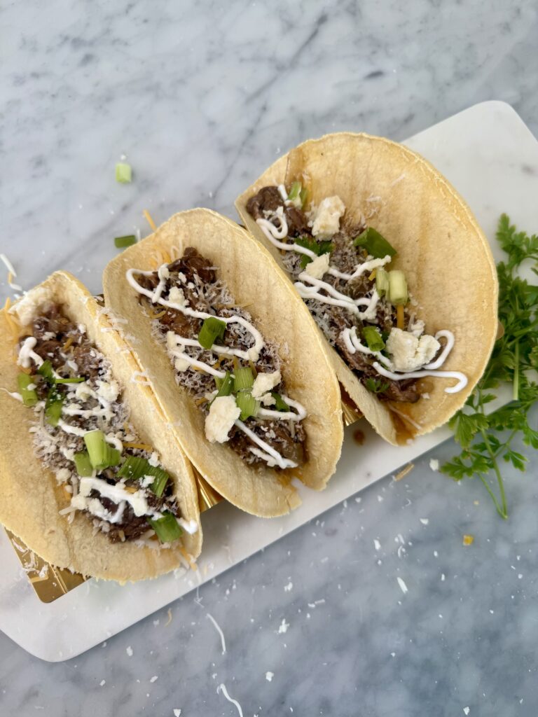 Three tacos filled with shredded meat, topped with shredded cheese, sour cream, green onions, and crumbled cheese, served on a white tray with parsley garnish on a marble surface.