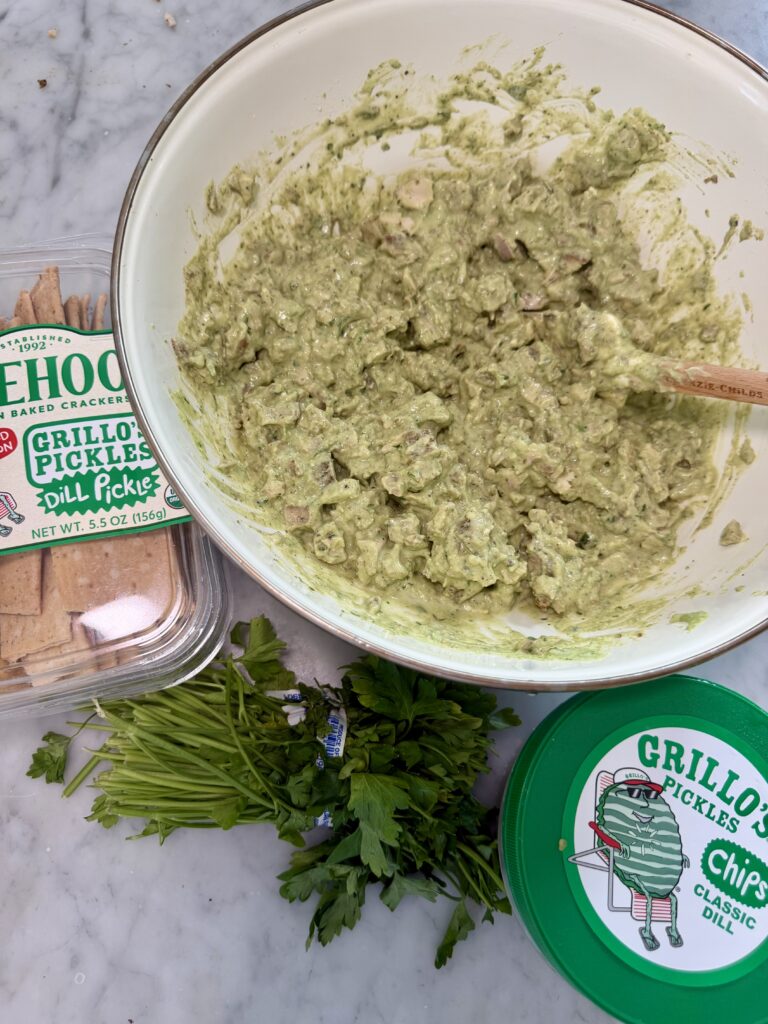A bowl of green dip with a spoon, surrounded by crackers, Grillos Pickles containers, and fresh parsley on a marble countertop—perfect for serving alongside your favorite Green Godess Chicken Salad or Grillo Chicken Salad.