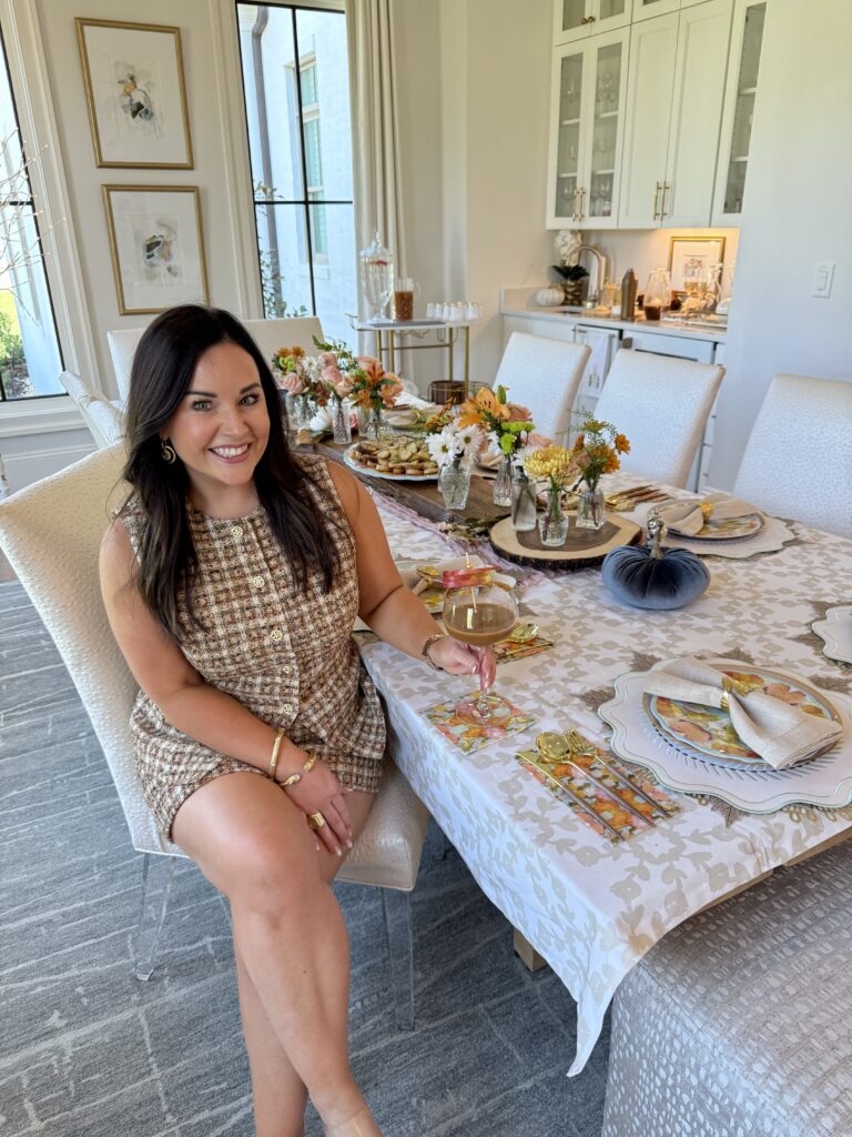 A woman in a tweed outfit sits at a dining table set with floral arrangements, food, and elegant place settings, smiling and holding a glass of wine in a bright, stylish dining room—a perfect scene to inspire your own party hosting tips.