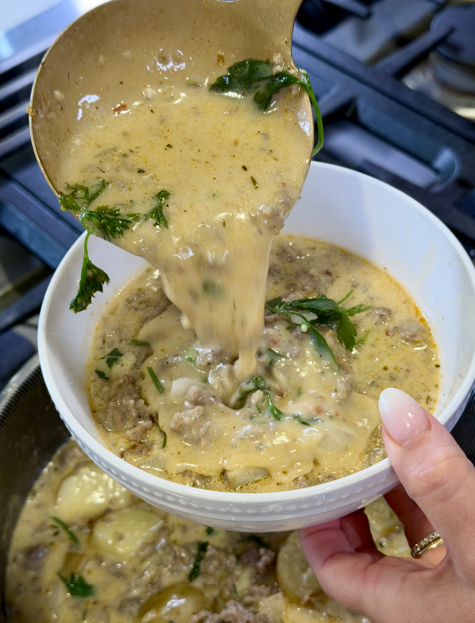 A hand holds a white bowl as creamy soup with ground meat, potatoes, and herbs is ladled in from a pot on a stove.