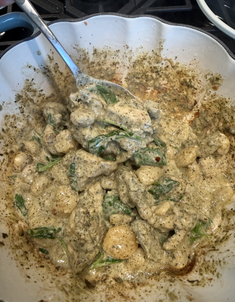 Creamy gnocchi with spinach and pesto sauce in a white scalloped bowl, being stirred with a large metal spoon.