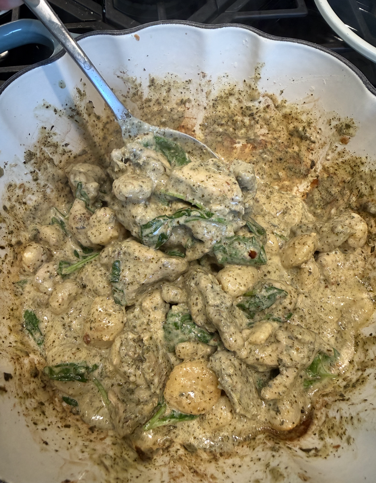 Creamy gnocchi with spinach and pesto sauce in a white scalloped bowl, being stirred with a large metal spoon.
