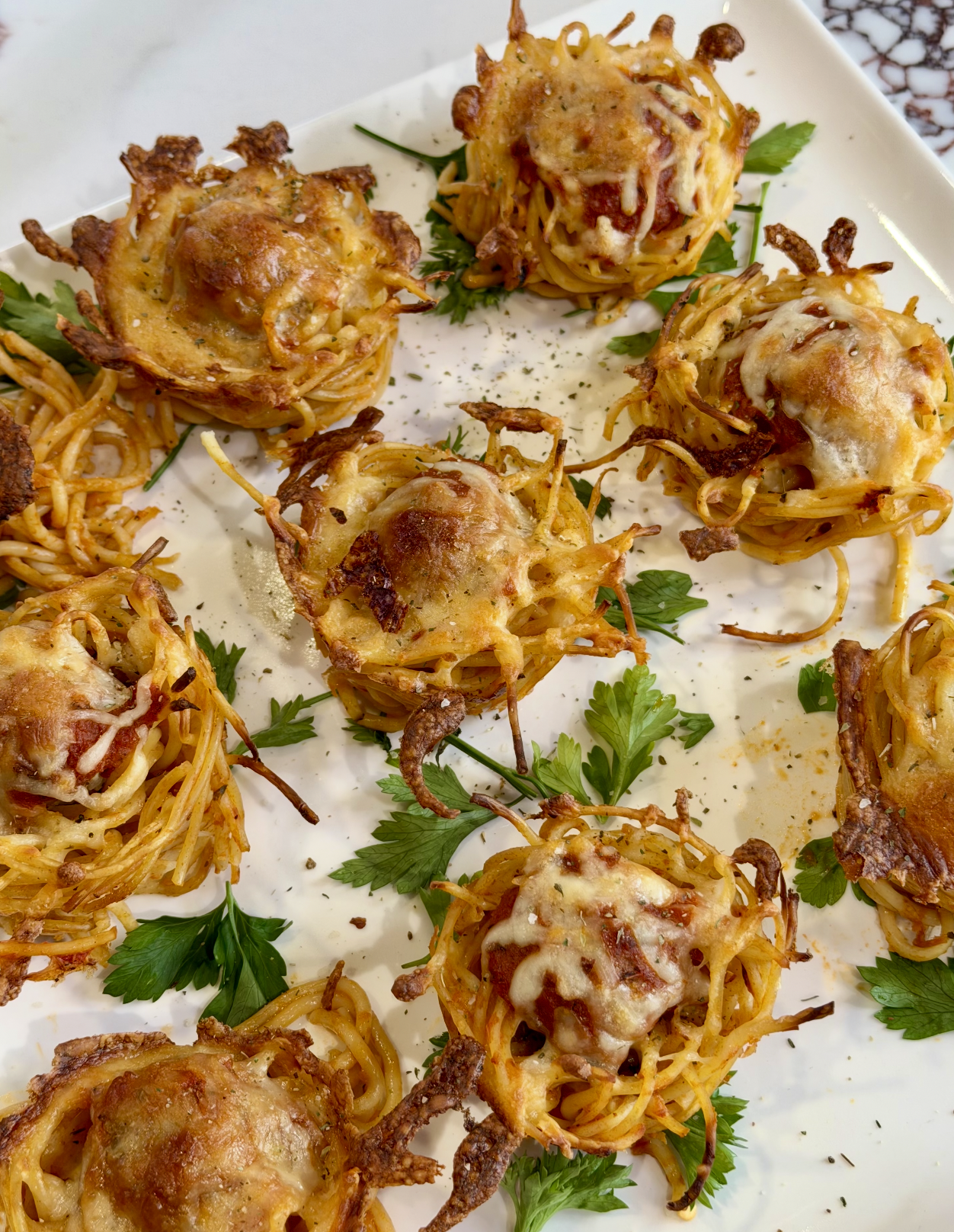 Baked spaghetti nests topped with melted cheese and herbs, each nest containing a meatball, arranged on a white platter garnished with fresh parsley.