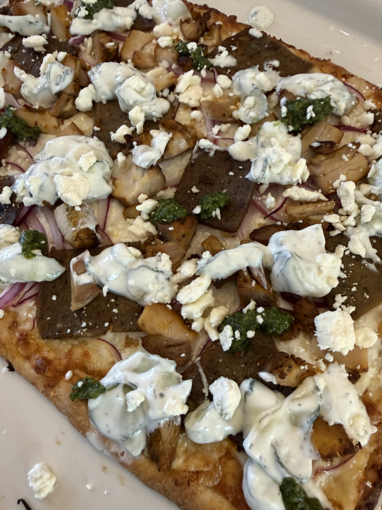 Flatbread pizza topped with chunks of chicken, gyro meat, sliced red onions, dollops of creamy white sauce, crumbled feta cheese, and green pesto on a golden-brown crust.