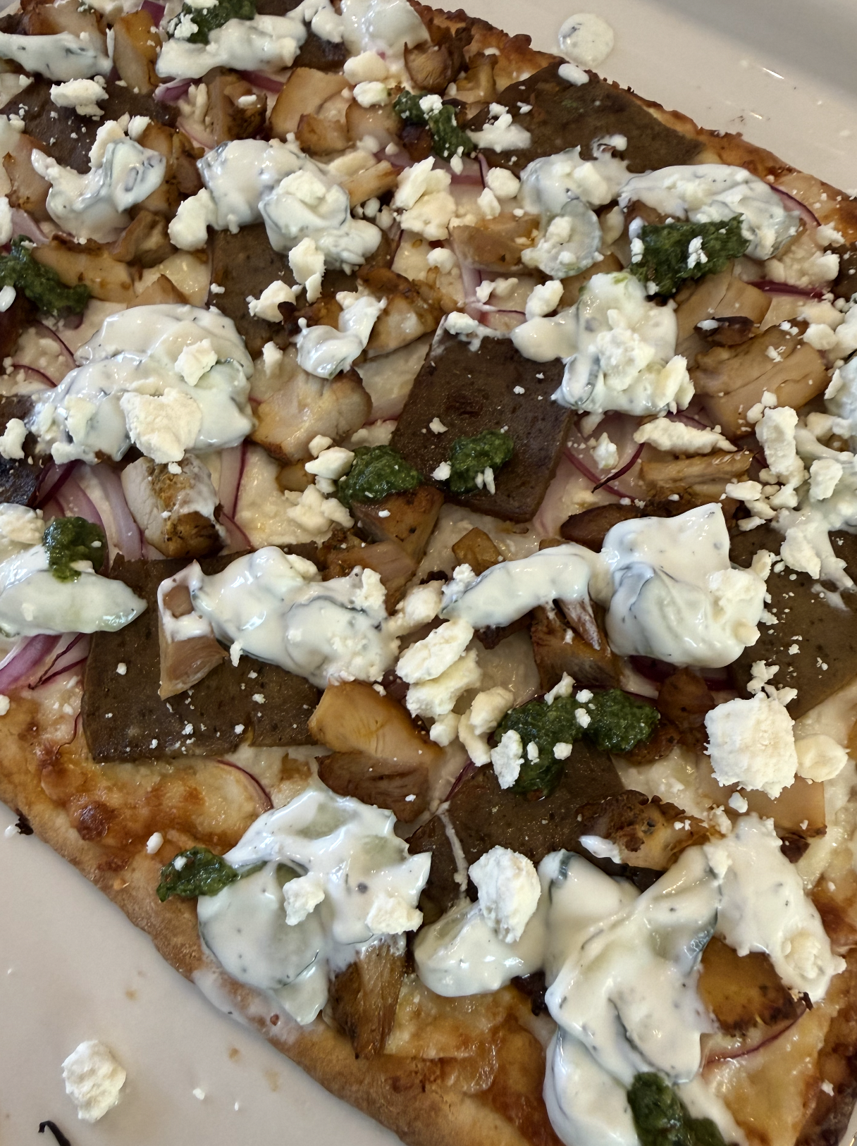 Flatbread pizza topped with chunks of chicken, gyro meat, sliced red onions, dollops of creamy white sauce, crumbled feta cheese, and green pesto on a golden-brown crust.