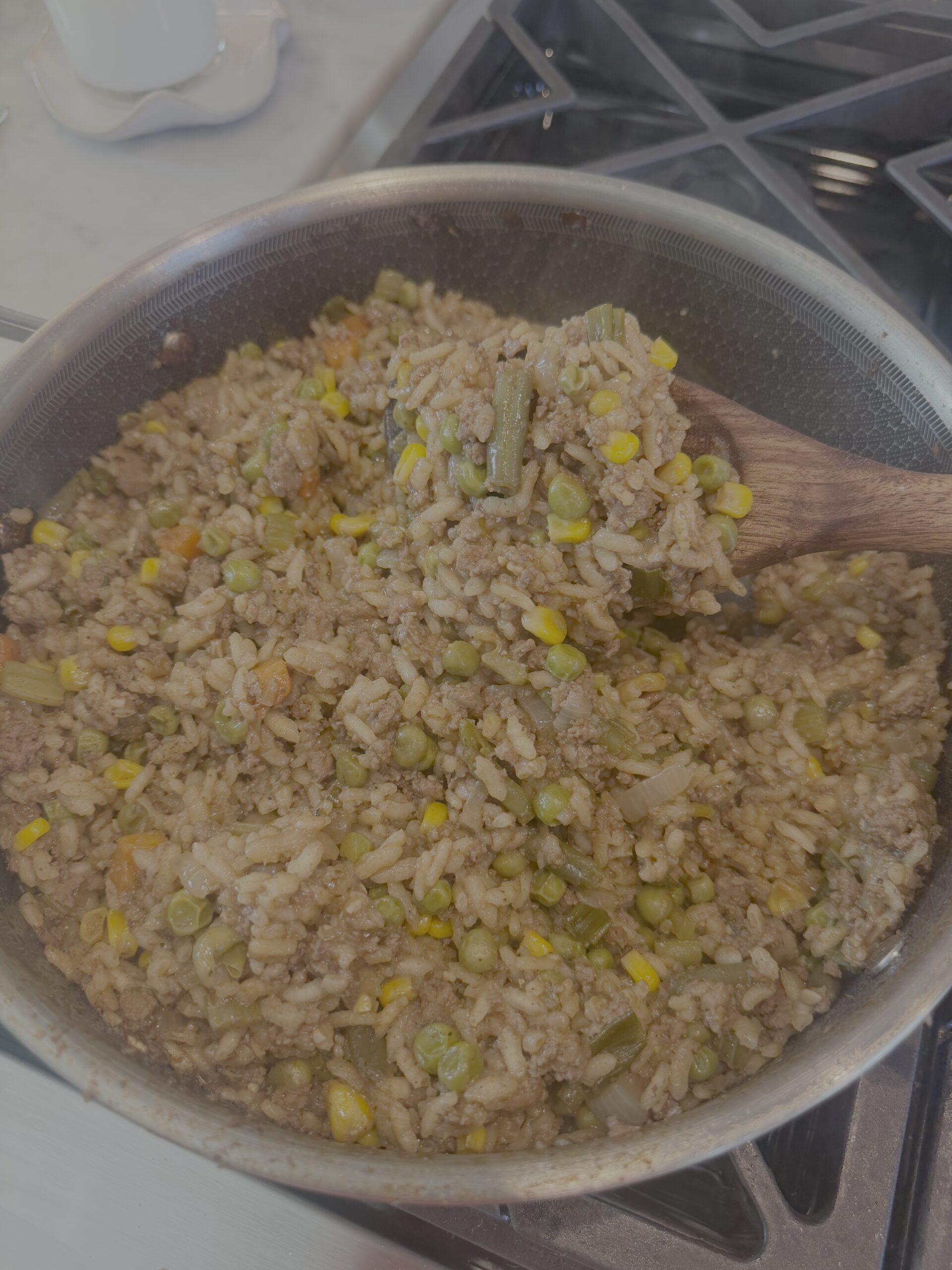 A pot of cooked ground beef mixed with rice, green beans, peas, corn, and carrots is being stirred with a wooden spoon on a stovetop.