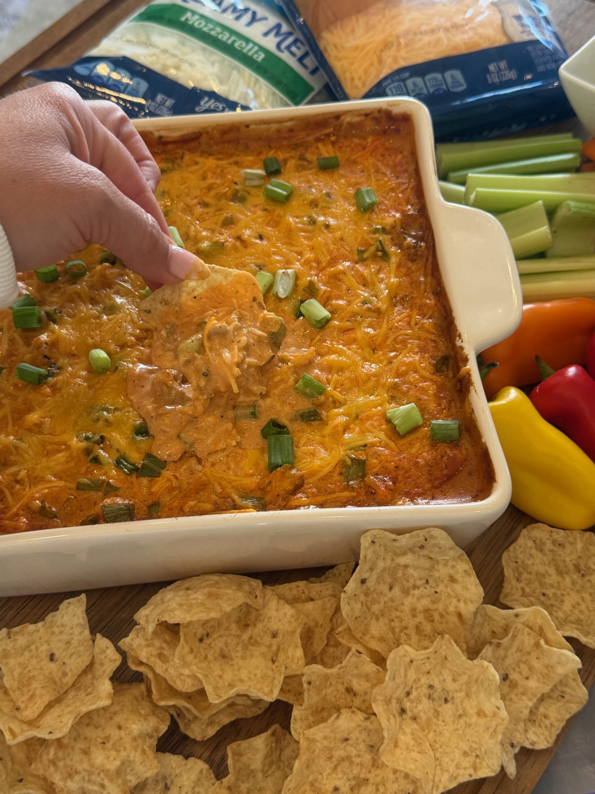 A hand dips a tortilla chip into a cheesy Buffalo Chicken Dip topped with green onions, surrounded by tortilla chips, celery, mini bell peppers, and packs of shredded cheese.