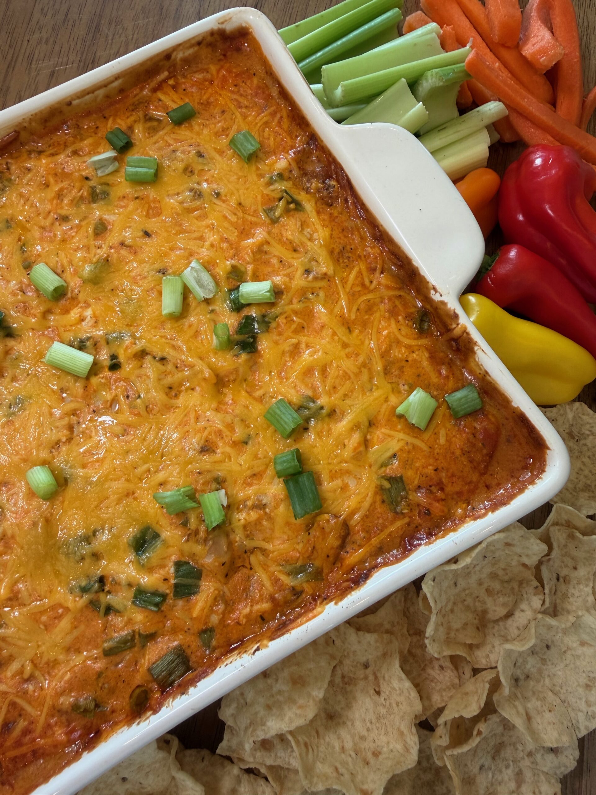 A baked cheesy Buffalo Chicken Dip topped with green onions in a white dish, surrounded by tortilla chips, celery, carrot sticks, and assorted bell pepper slices on a wooden table.
