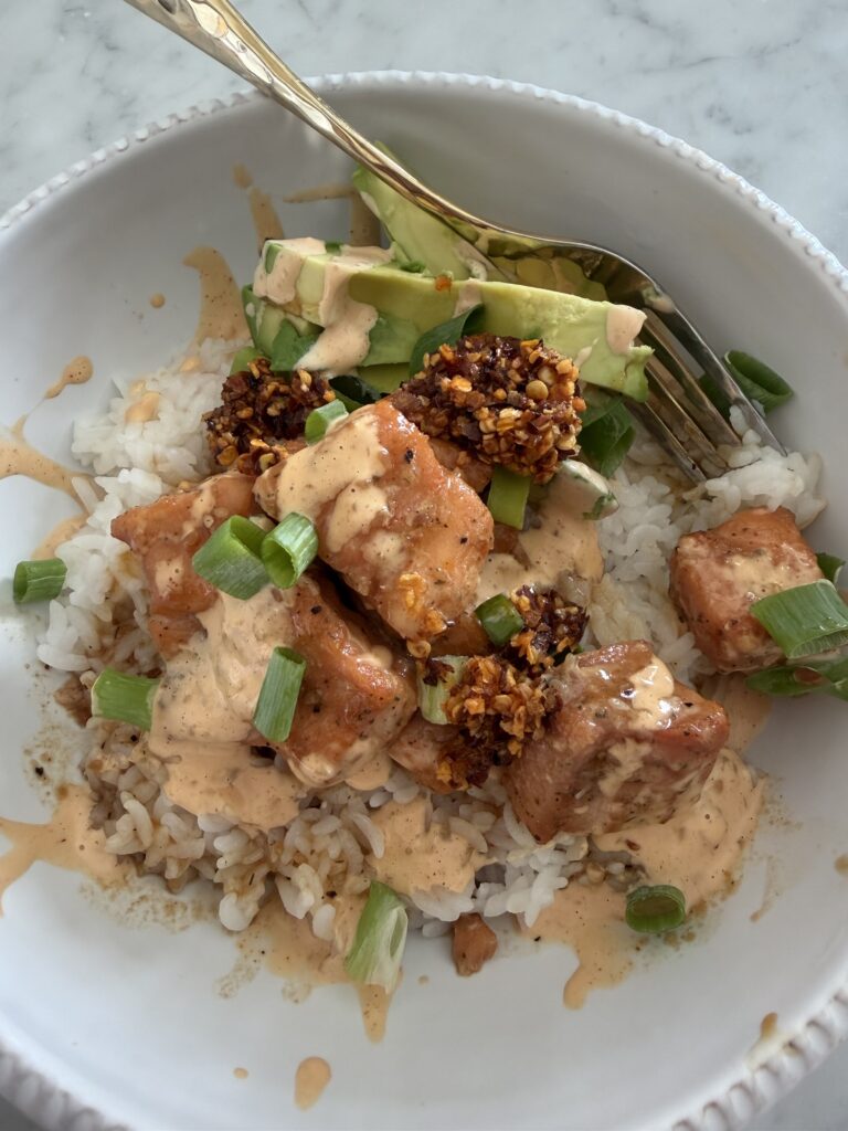 A white bowl with rice, seared salmon cubes, sliced avocado, chopped green onions, chili crisp, and creamy orange sauce, with a fork on the side.