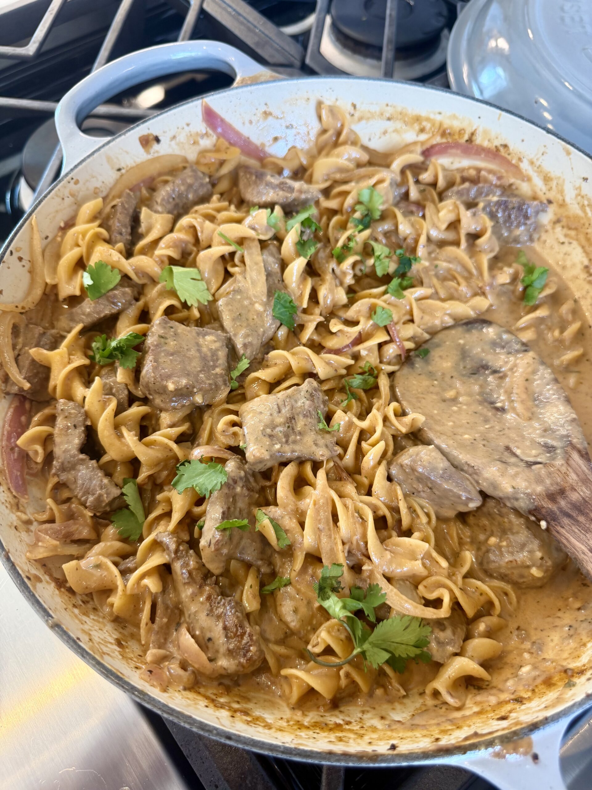 A skillet filled with creamy beef stroganoff, featuring wide egg noodles, sliced beef, sauce, and fresh parsley garnish, with a wooden spatula resting in the pan.