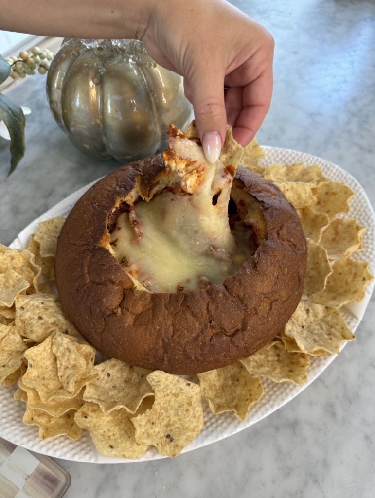 A hand dips a tortilla chip into a bread bowl filled with melted cheesy dip, surrounded by more chips on a white platter. A decorative pumpkin sits in the background.
