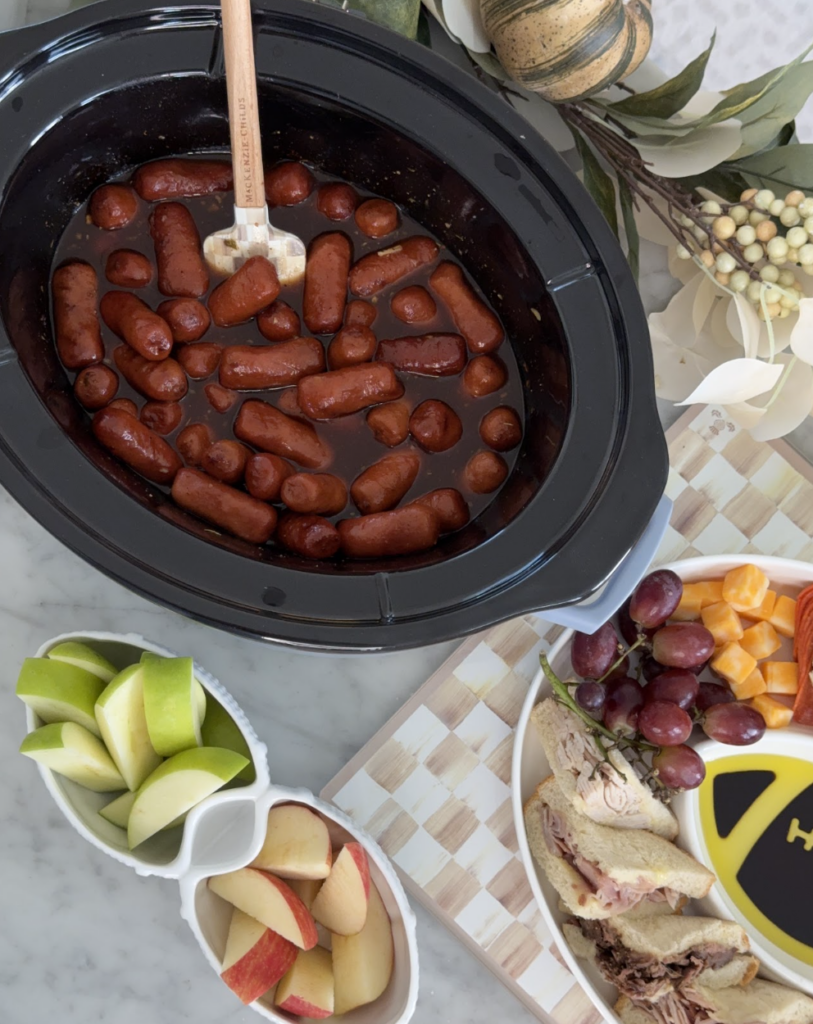 A slow cooker filled with cocktail sausages in sauce sits on a counter next to plates of apple slices, grapes, and sandwiches. A spatula rests in the slow cooker.