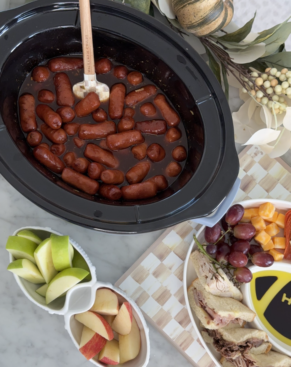 A slow cooker filled with cocktail sausages in sauce sits on a counter next to plates of apple slices, grapes, and sandwiches. A spatula rests in the slow cooker.