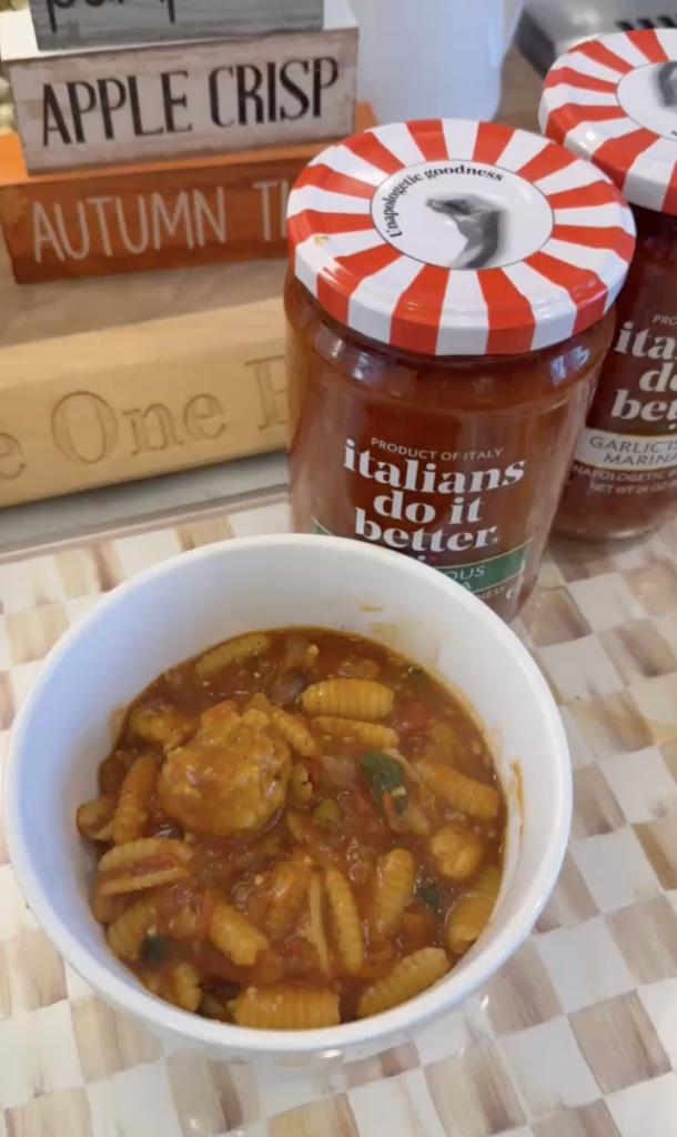 A white bowl of cavatelli pasta with tomato sauce and chicken meatballs sits on a woven placemat, next to a jar labeled italians do it better. Decorative autumn-themed wooden blocks are in the background.