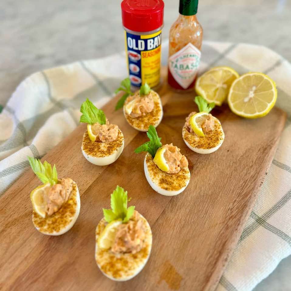Deviled eggs topped with lemon slices, celery, and seasoning on a wooden board, with Old Bay, Tabasco sauce, and lemon halves in the background. A checkered cloth is partially visible under the board.