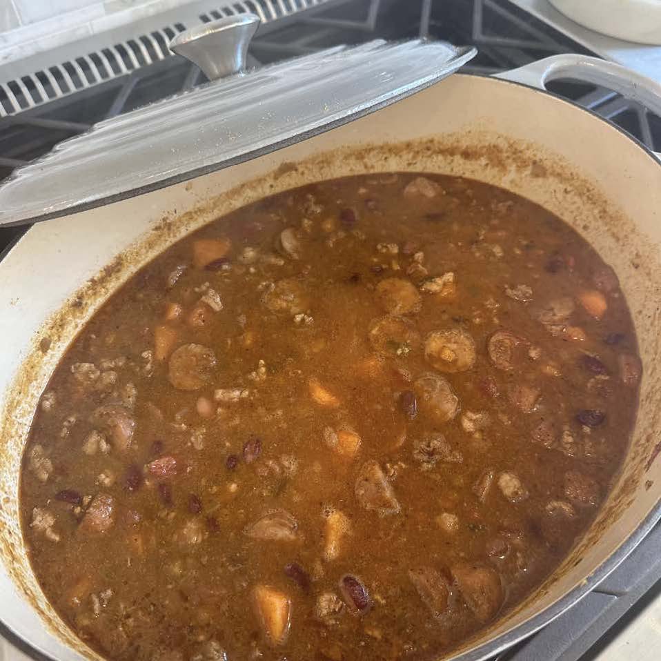 A large pot of chili with beans, vegetables, and meat simmers on a stove, partially covered by a lid.