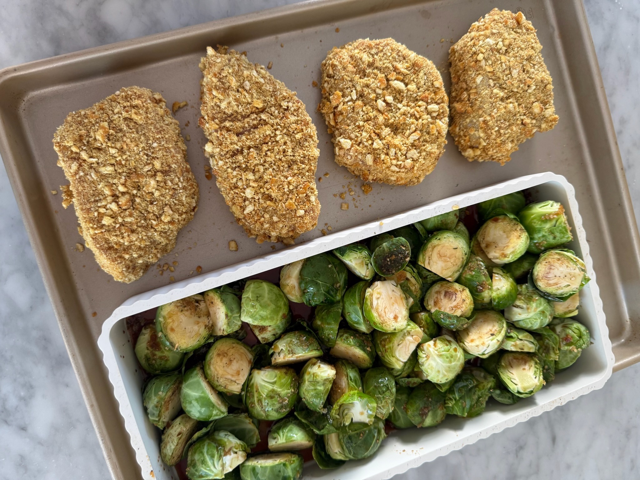 A baking sheet with four breaded chicken breasts and a tray filled with halved Brussels sprouts, ready to be cooked, on a marble countertop.