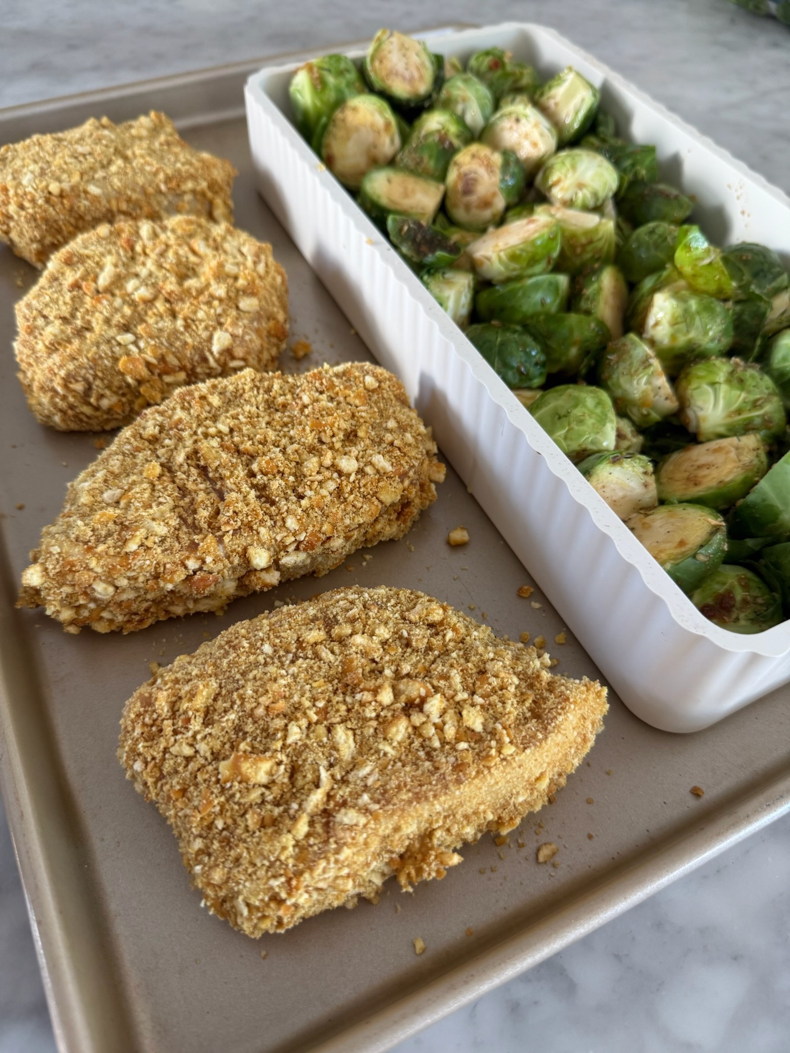 Four breaded chicken breasts on a baking sheet next to a white dish filled with seasoned, halved Brussels sprouts, ready to be cooked.