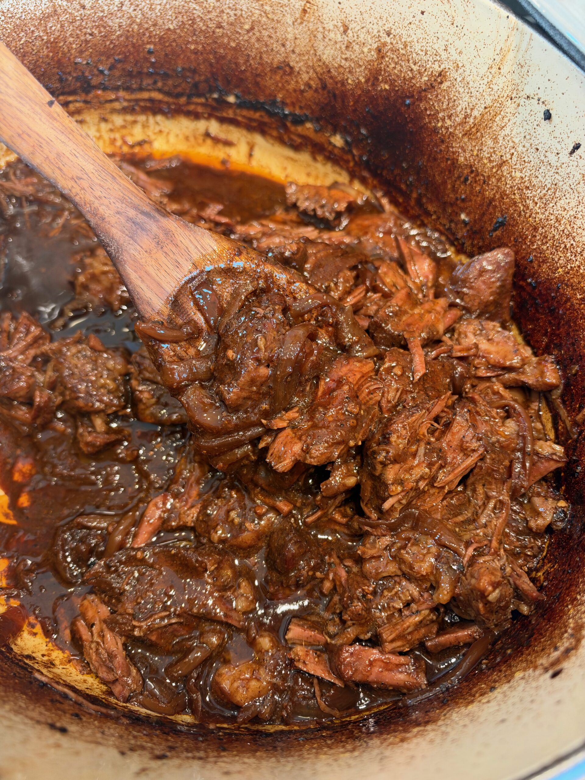 A wooden spoon lifting tender shredded French onion pot roast and caramelized onions from a Dutch oven.