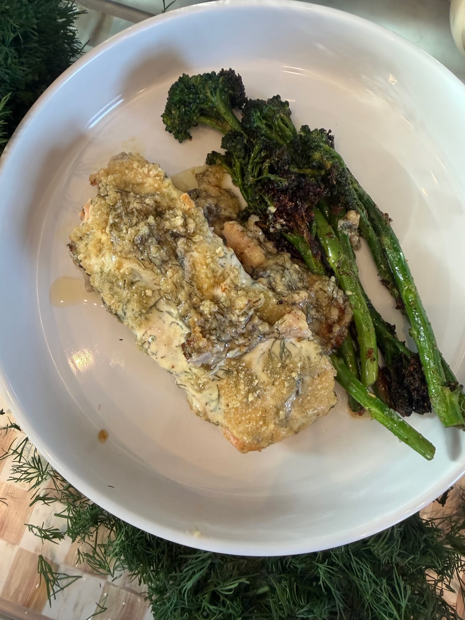 A fillet of creamy lemon dill Parmesan salmon with a golden topping, served with charred broccolini on a white plate.