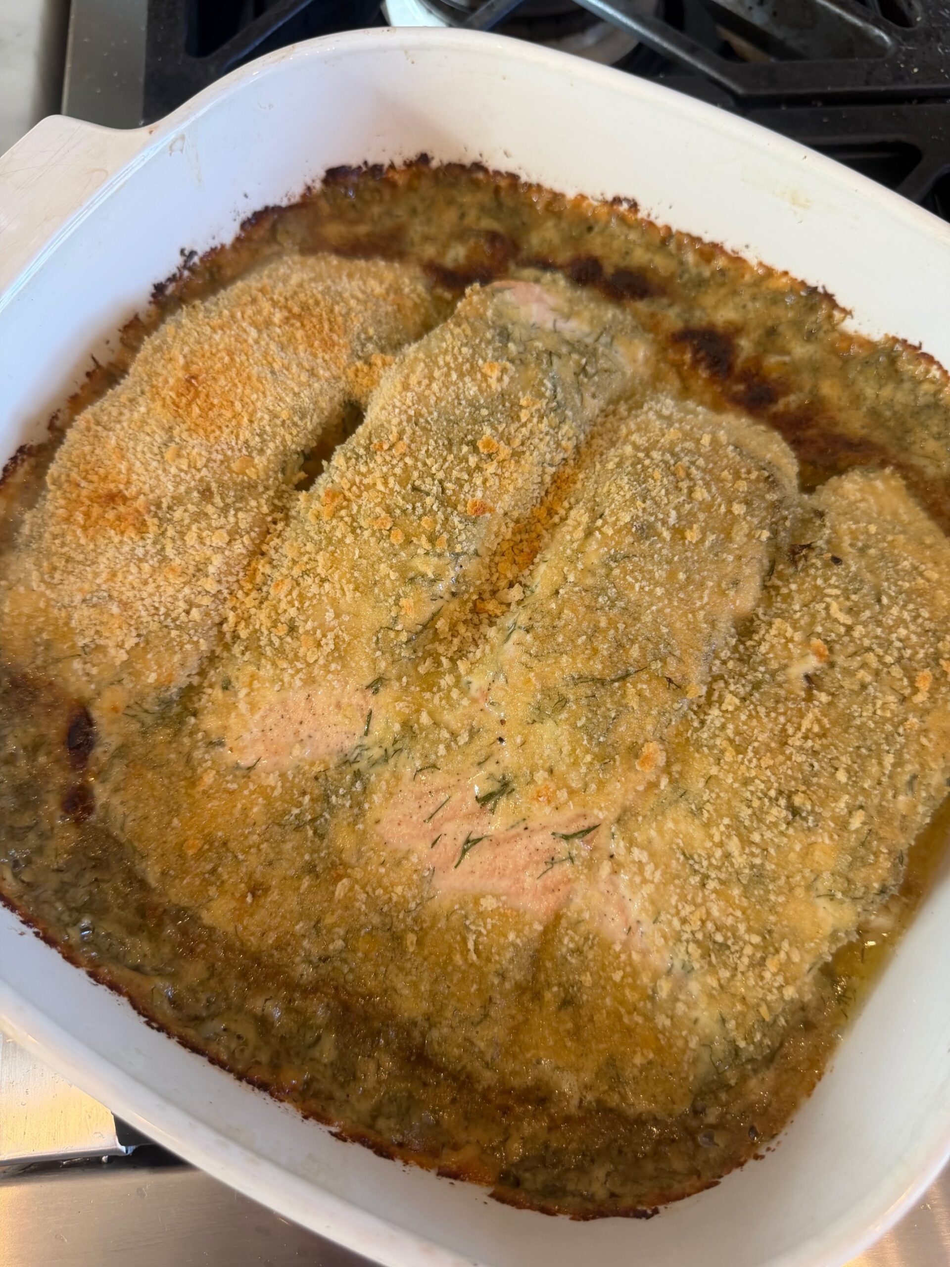 Four lemon dill salmon fillets with a golden, bubbly Parmesan and fresh dill topping in a white ceramic baking dish.