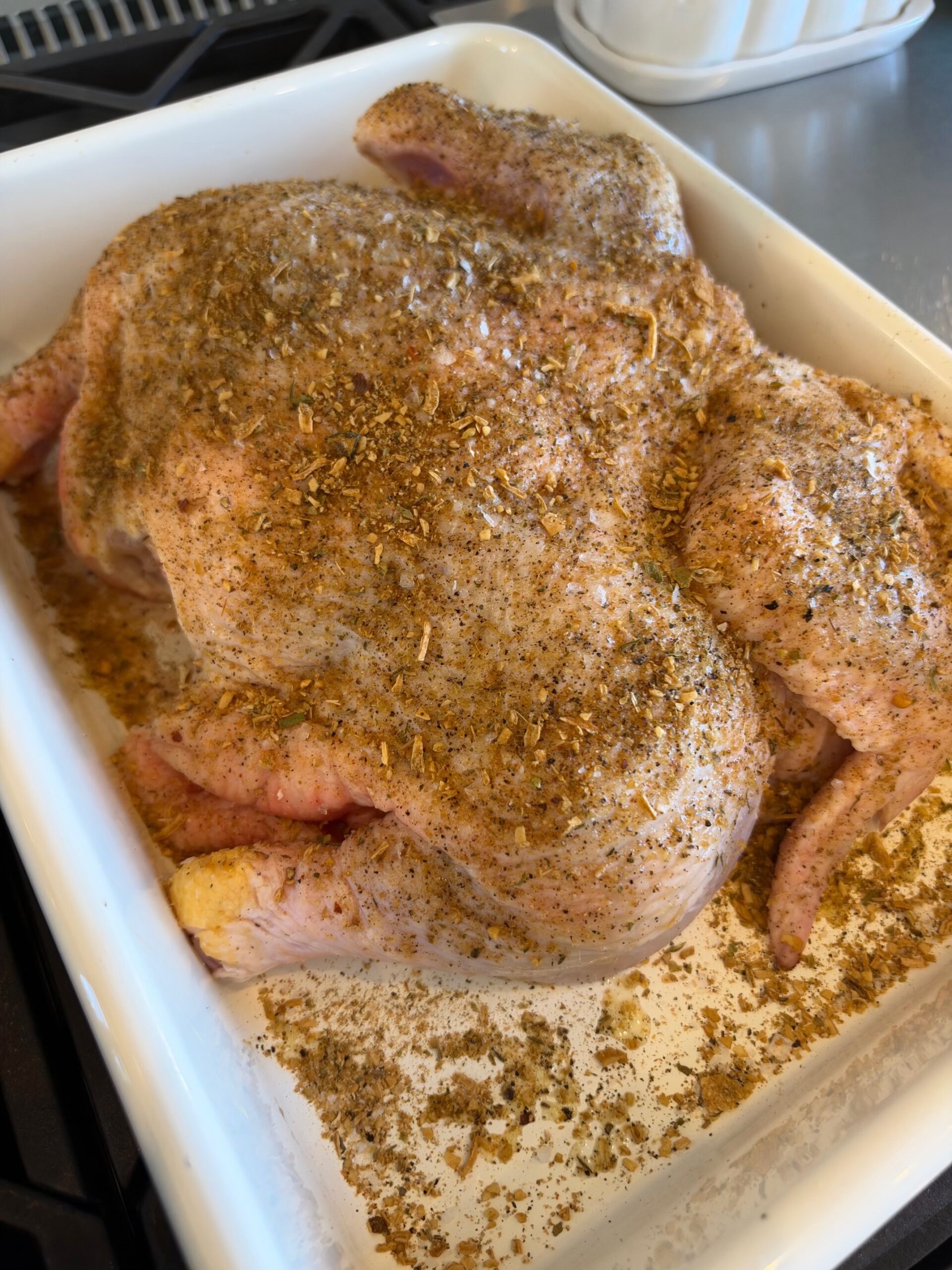 A spatchcocked whole chicken seasoned with Creole Trinity & Herb seasoning and black pepper in a white baking dish, ready to roast.