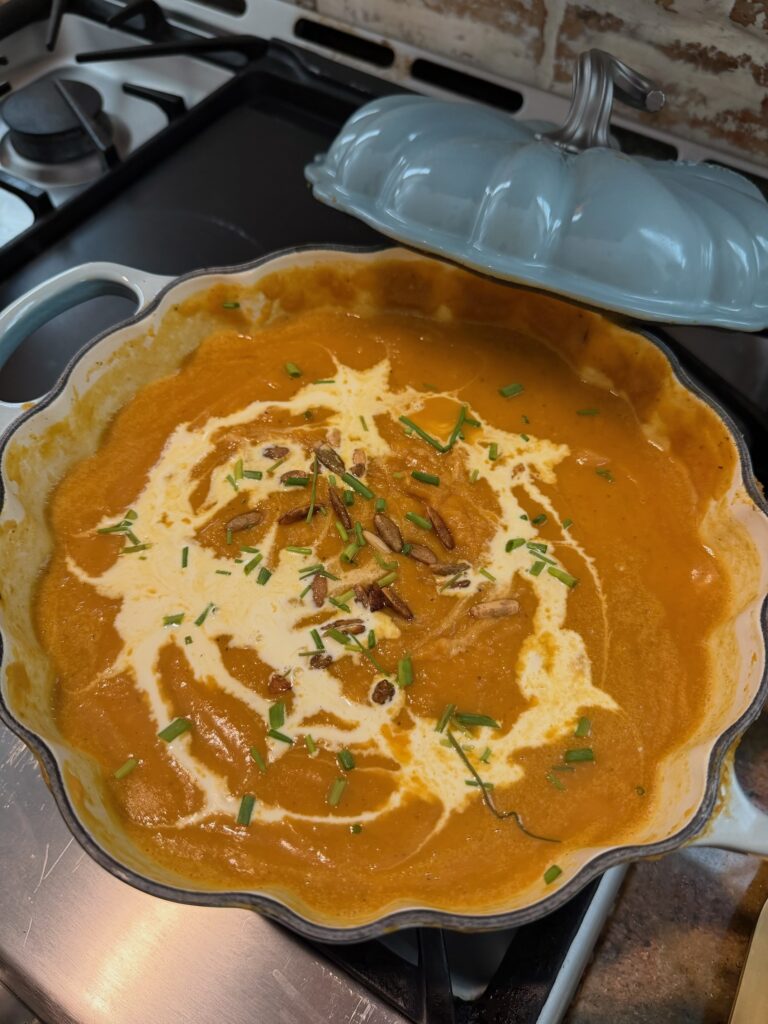 Creamy roasted butternut squash and sweet potato soup in a blue braiser, topped with a swirl of cream, toasted pepitas, and fresh herbs.